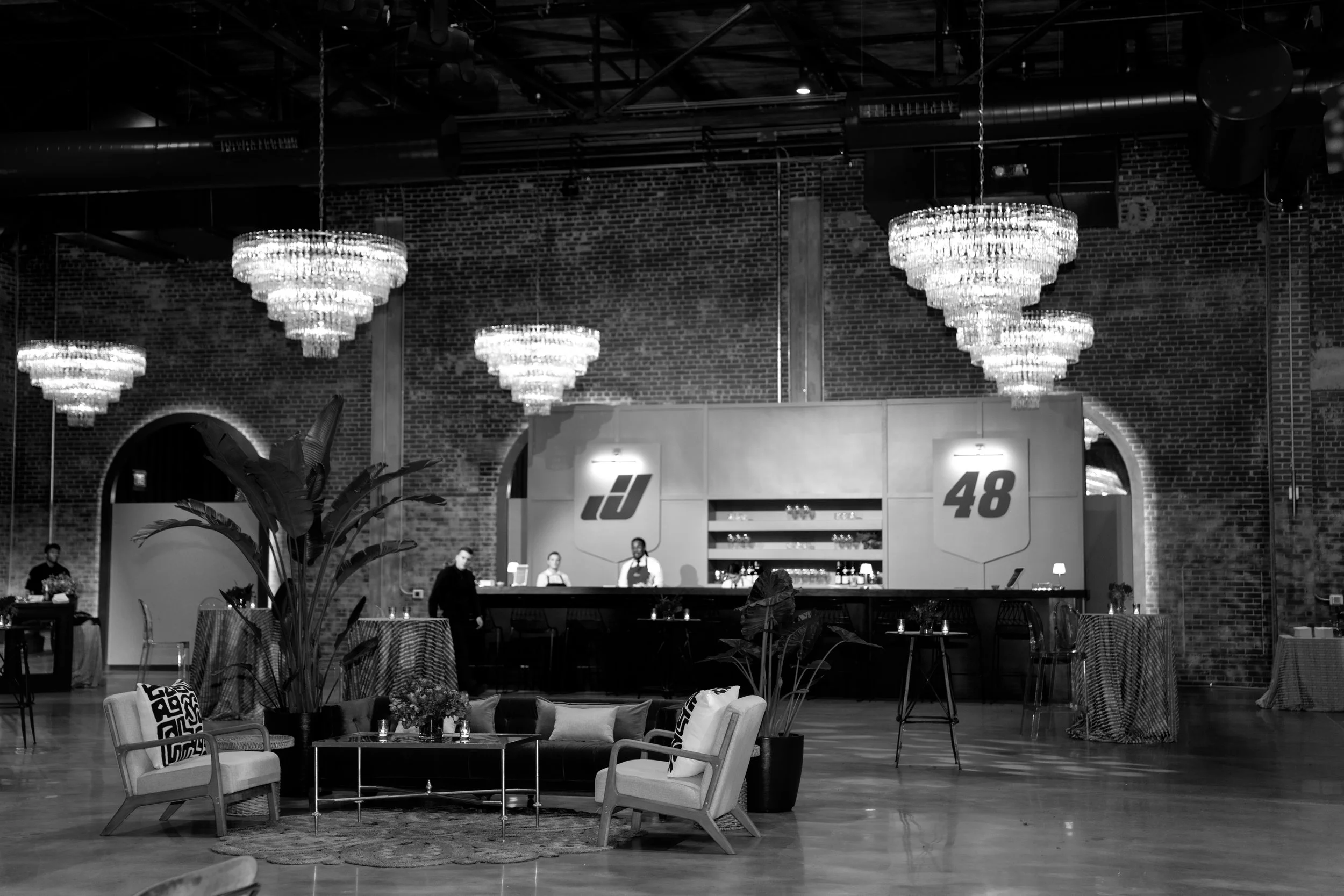 A black and white photo of a modern event space with chandeliers, decorative brick walls, and a bar counter. There are seating areas with sofas and tables.