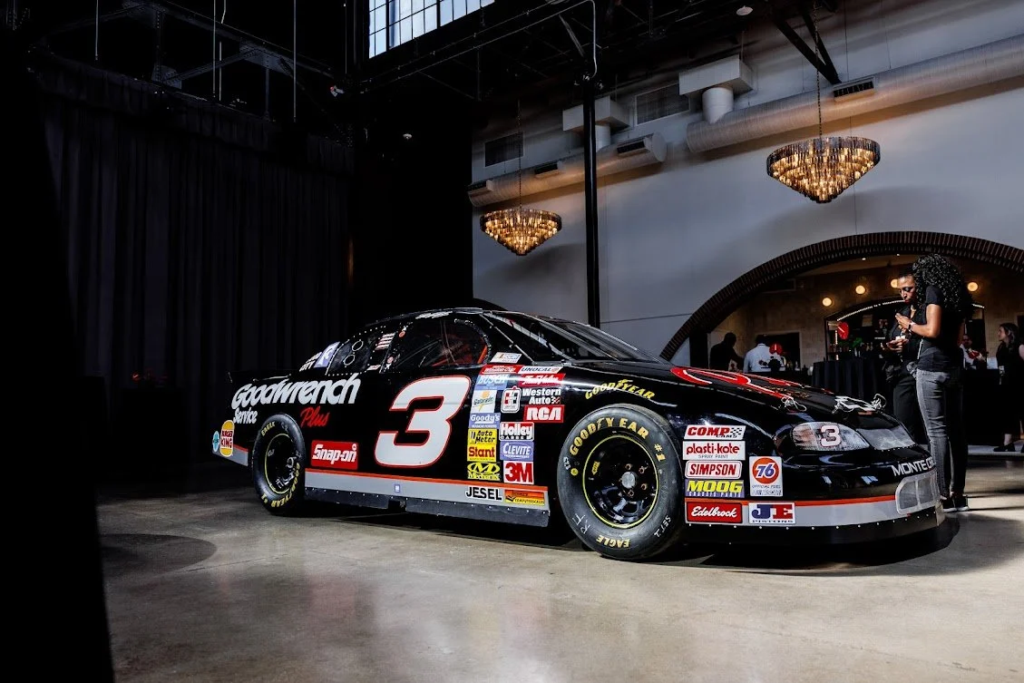 Black race car with the number 3 and sponsor logos, displayed indoors at an event, with a woman standing beside it using her phone.