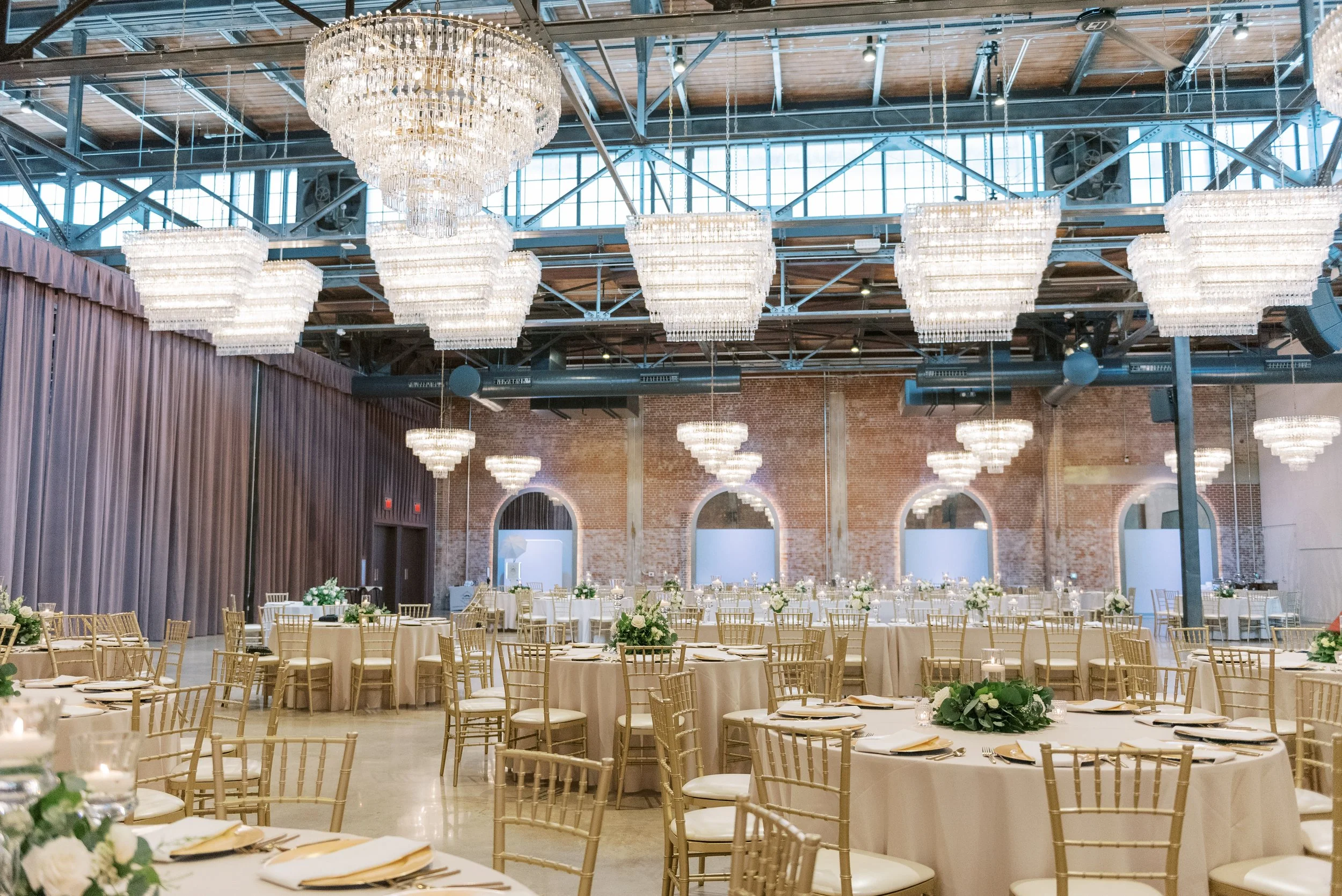 Elegant banquet hall decorated with chandeliers, round tables with white tablecloths and floral centerpieces, set for a formal event in a brick-walled industrial-style venue.
