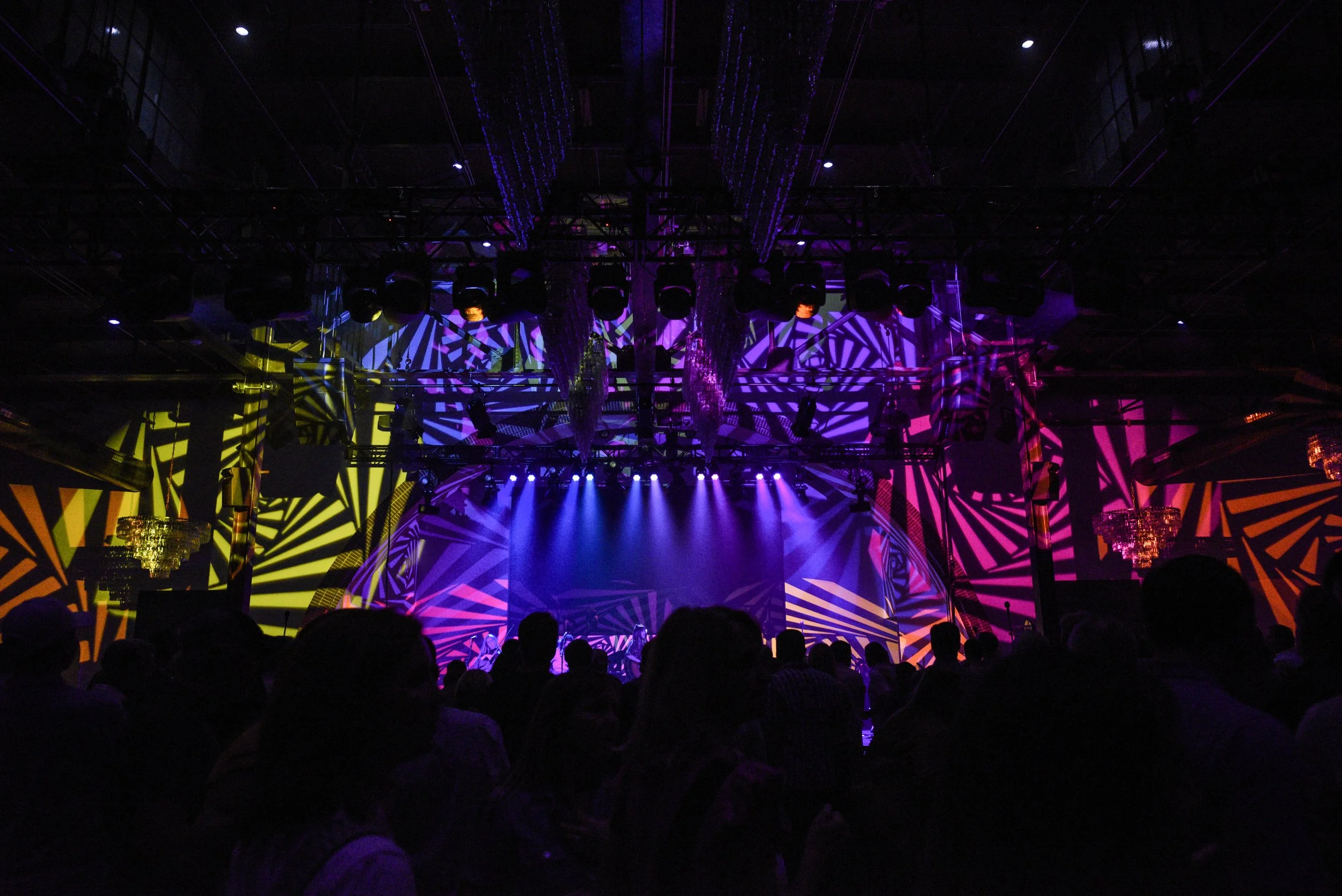 A concert or event stage with vibrant purple, yellow, pink, and blue geometric lighting projections, with an audience in silhouette.