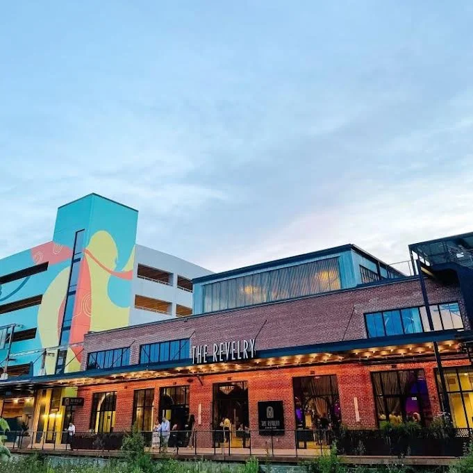 Exterior of a modern building called 'The Revelry' with a brick facade and large windows, featuring a colorful mural on an adjacent building, evening sky above, people walking outside.
