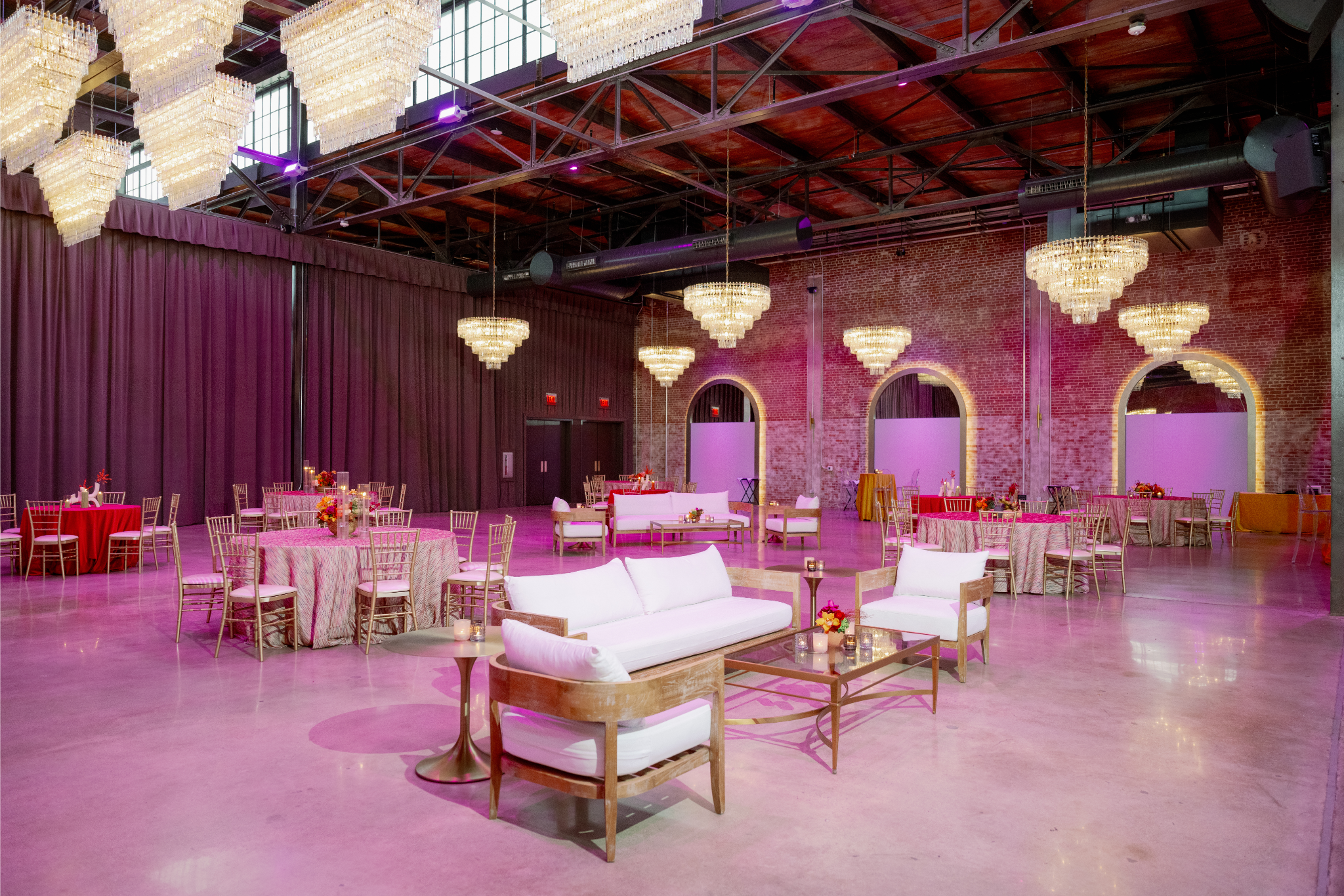 Large event space decorated with chandeliers, round tables with pink tablecloths, and white sofas with wooden frames. The space has exposed brick walls, large arched windows covered with purple curtains, and a polished concrete floor.