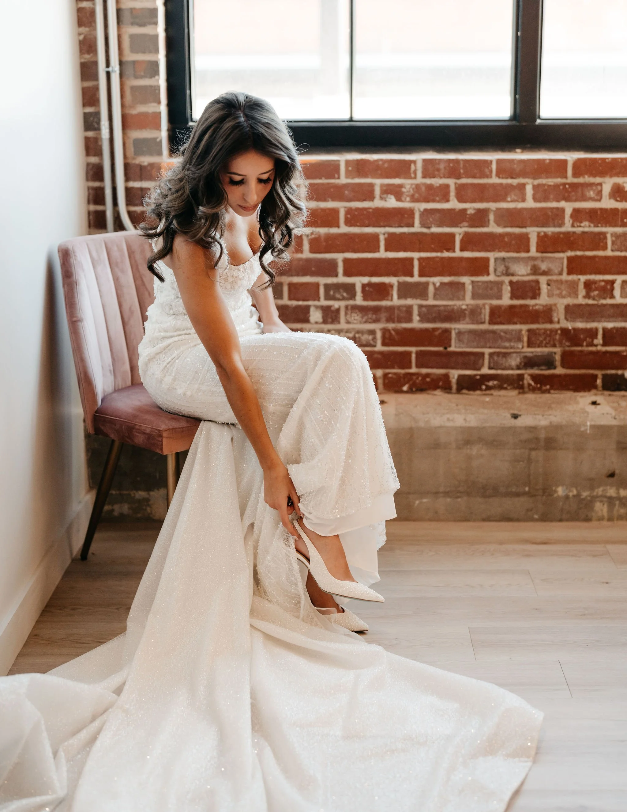 A bride in a white wedding dress putting on her high heels next to a brick wall and a window.