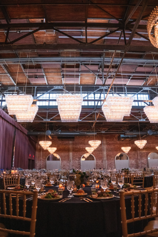Empty banquet hall decorated for an event with round tables set with glassware, plates, and floral centerpieces, chandeliers hanging from a high wooden ceiling, and brick walls with arched windows.