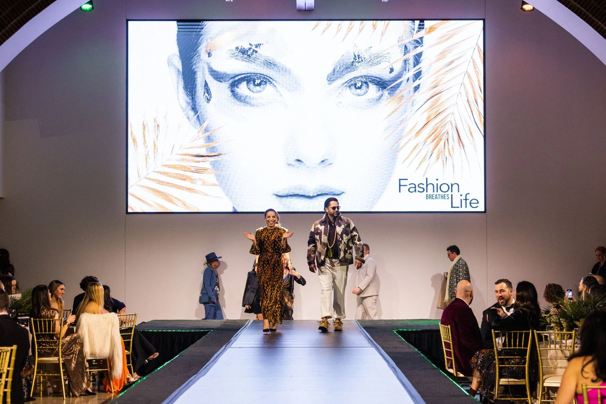 Models walking down runway at a fashion show, large screen displaying a model's face with the text 'Fashion Breaths Life' in the background