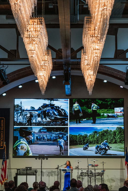 Large digital screen displaying four images related to emergency response and law enforcement, with a woman speaking at a podium in front of an audience in a conference hall.