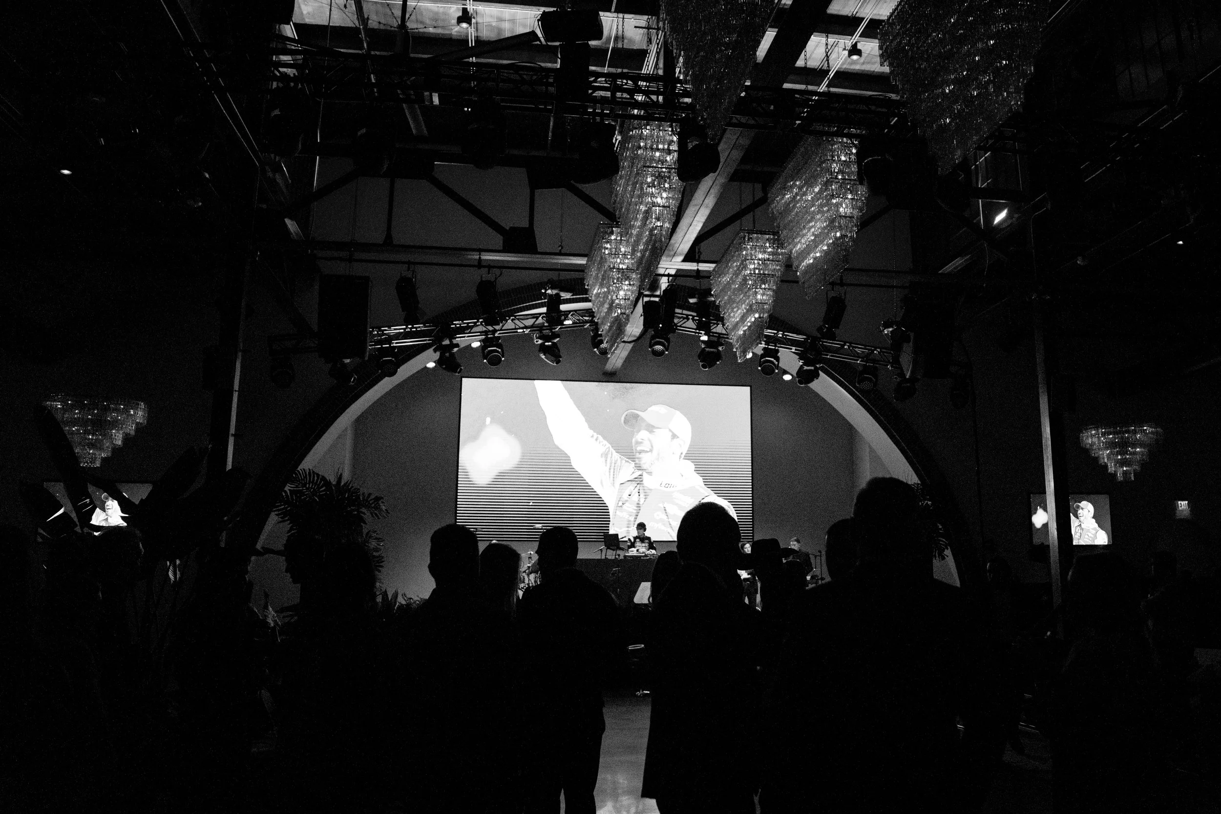 Black and white photo of a concert or event with people facing a large screen showing a performer with a cap, as well as a person in front of the screen. The setting is indoors with chandeliers and stage lighting.