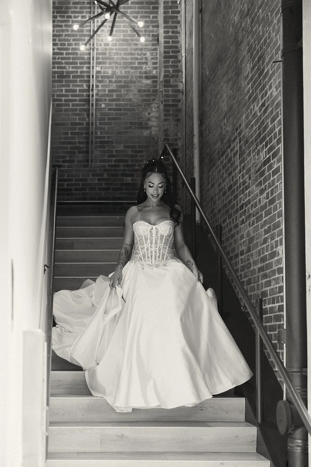 A woman in a strapless wedding gown with lace bodice and a full skirt walking down a staircase with brick walls and modern lighting.