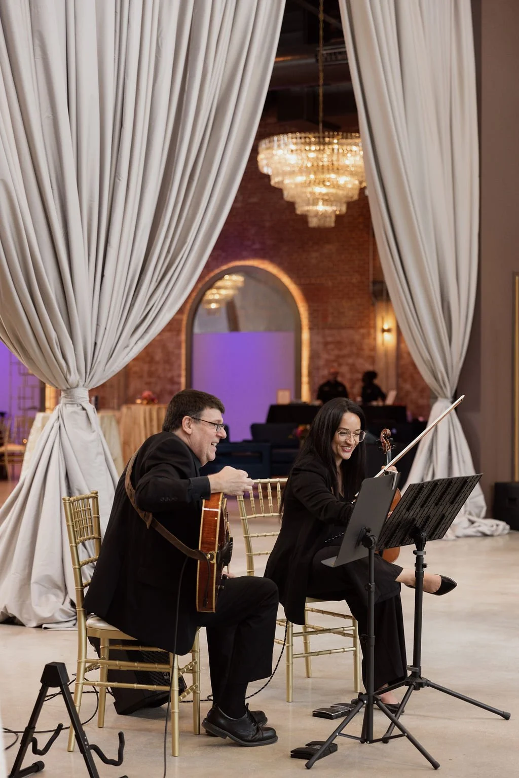 Two musicians, a man playing guitar and a woman playing violin, perform together in an elegant event space with draped curtains and a chandelier overhead.
