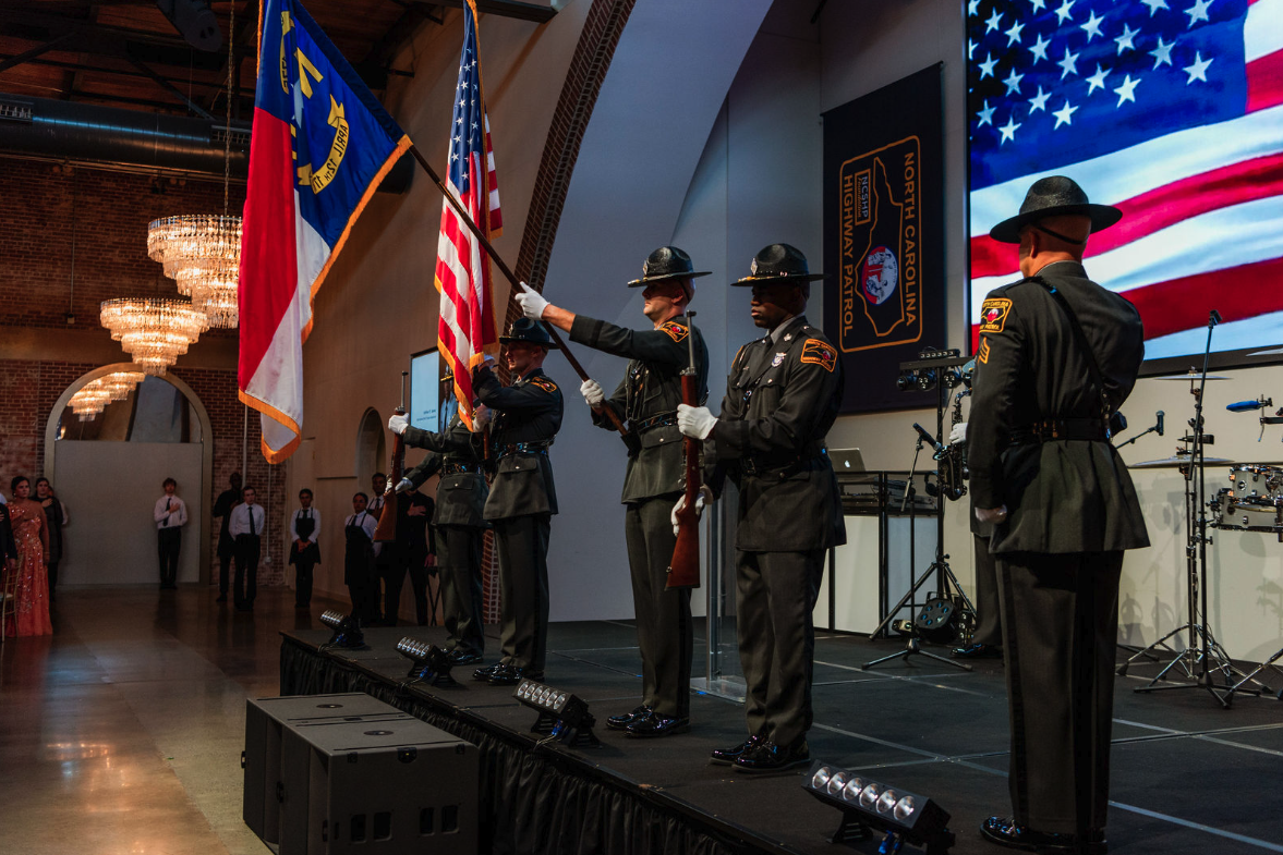 Law enforcement officers in formal uniform on stage during a ceremony, holding flags with a large American flag displayed in the background.