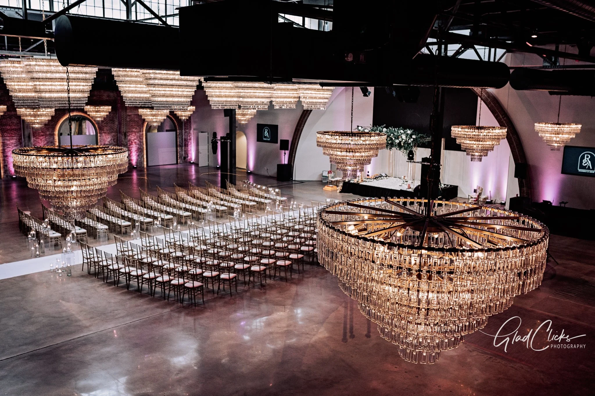 Elegant event space decorated with large chandeliers hanging over a stage area with floral arrangements, rows of chairs arranged for a ceremony, and purple uplighting on brick walls.