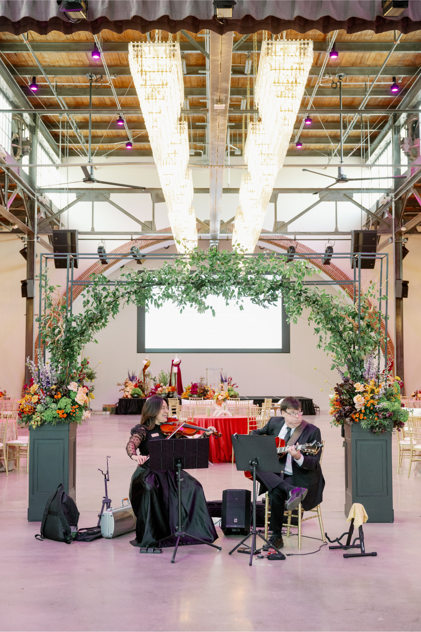 Two musicians performing with a violin and guitar at a decorated event space with floral arrangements and a large screen in the background.