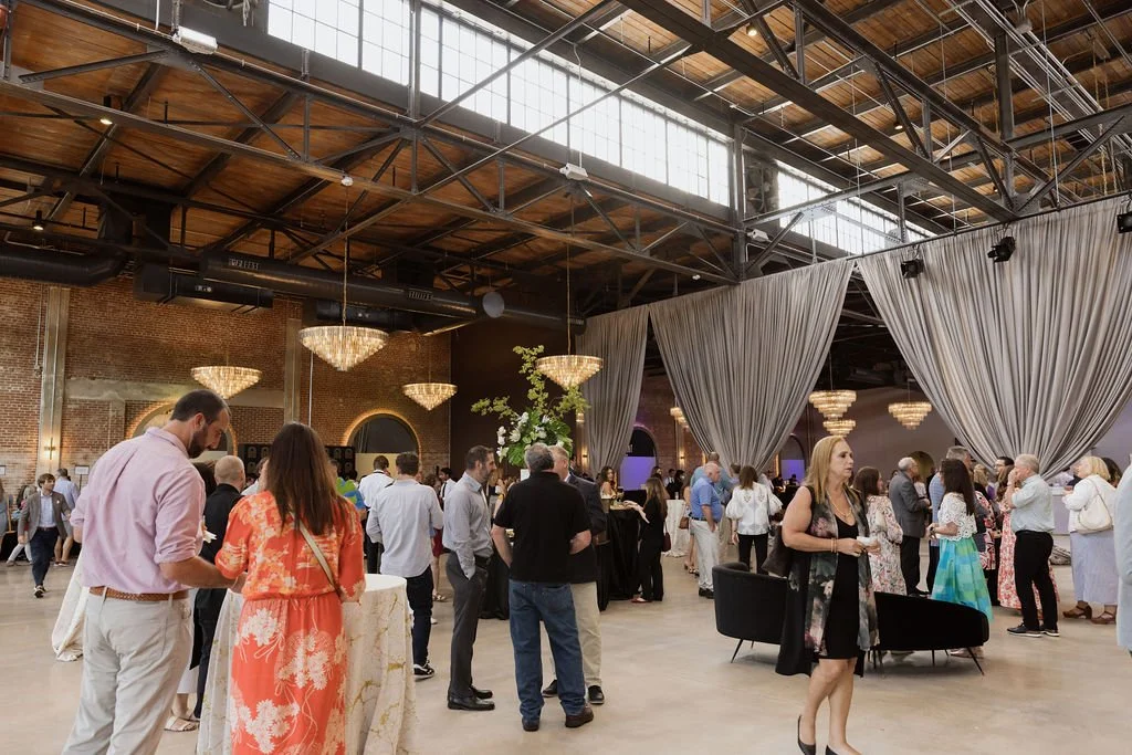 People gathered in a spacious event hall with high ceilings, large chandeliers, and draped curtains, attending a social or networking event.