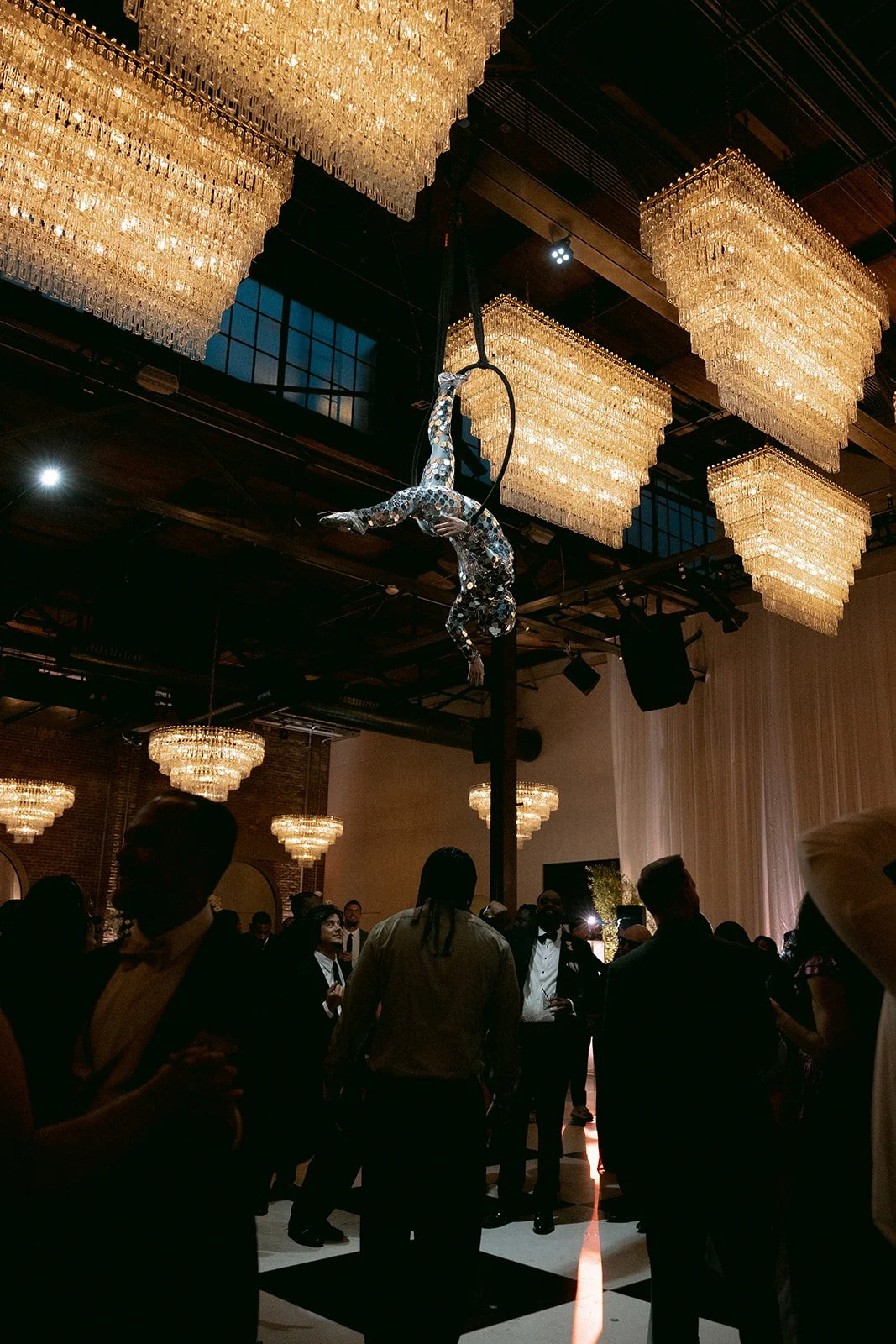 An aerial performer dressed in a sequined costume hangs from a trapeze in a ballroom decorated with large chandeliers, with guests in formal attire watching.