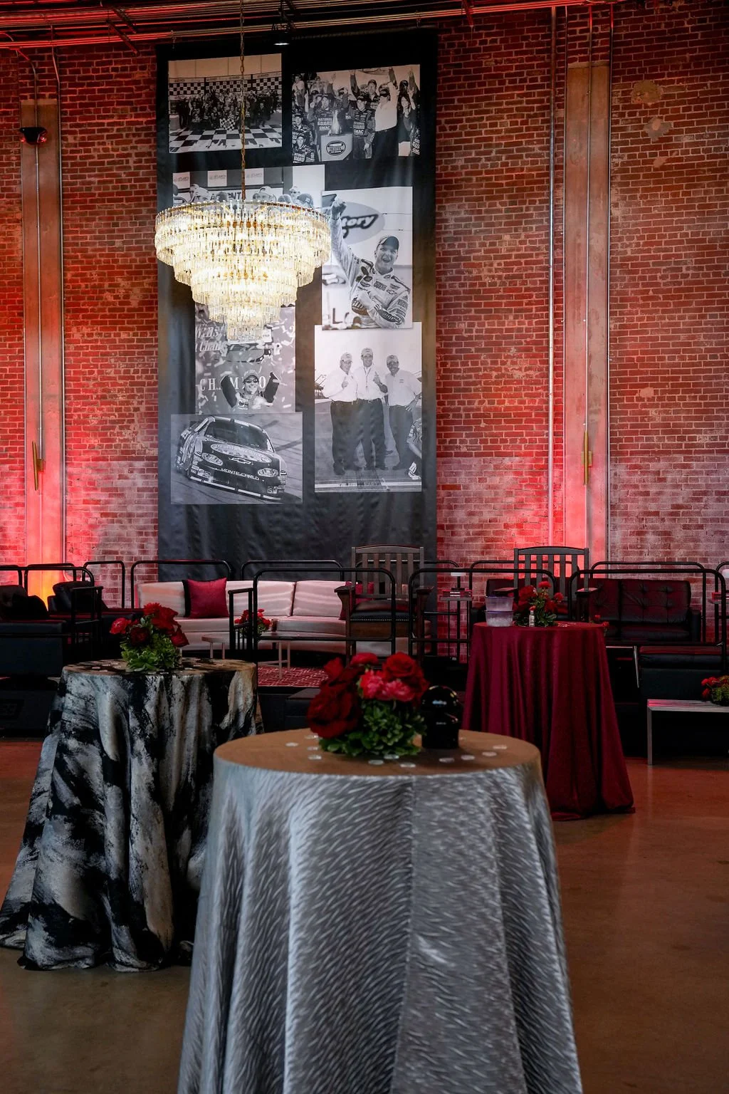 A decorated event space with round tables covered in different patterned tablecloths, red floral centerpieces, black chairs, a white sofa with red pillows, brick walls, a large black-and-white photo collage on fabric hanging from the ceiling, and a c