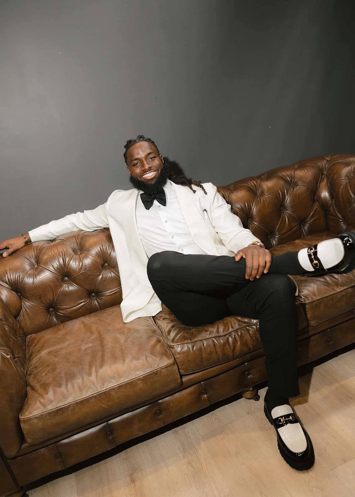 Man sitting on a brown leather couch, wearing a white tuxedo jacket, black pants, white shirt, black bow tie, and distinctive white and black shoes, smiling at the camera.