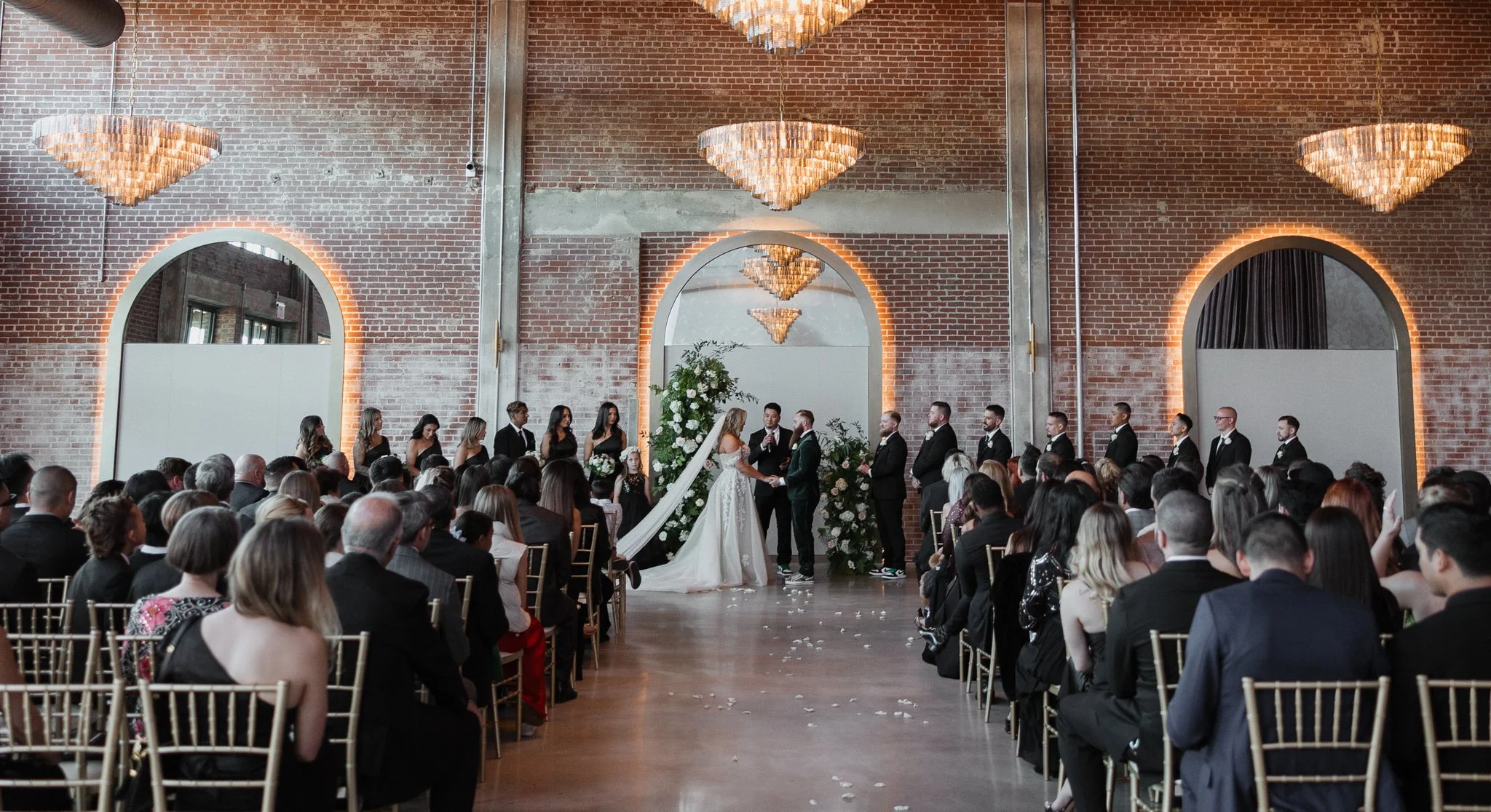 A wedding ceremony taking place in a modern industrial-style venue with brick walls and large arched mirrors. The bride and groom are exchanging vows at the altar, surrounded by their wedding party and seated guests.