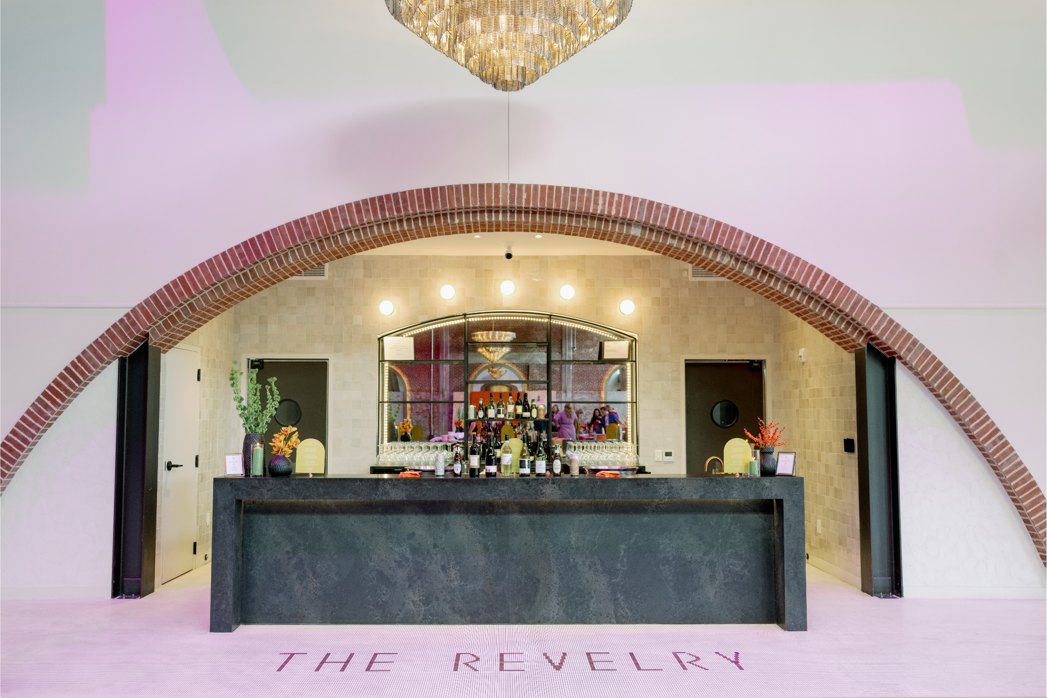 An elegant hotel or lounge reception area with a black marble counter, bottles of wine, and glasses. Behind the counter is a large mirror with a curved top, surrounded by a brick arch, with a chandelier hanging from the ceiling. The floor has pink ca