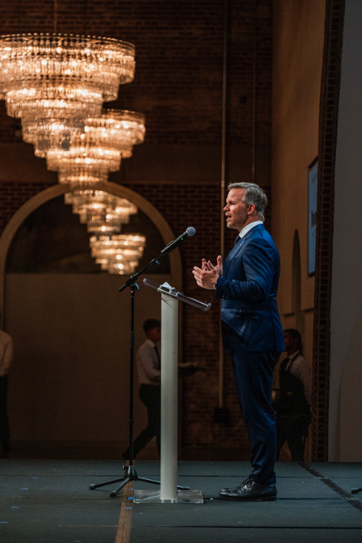 A man in a blue suit giving a speech at a podium on a stage with chandeliers hanging from the ceiling in the background.