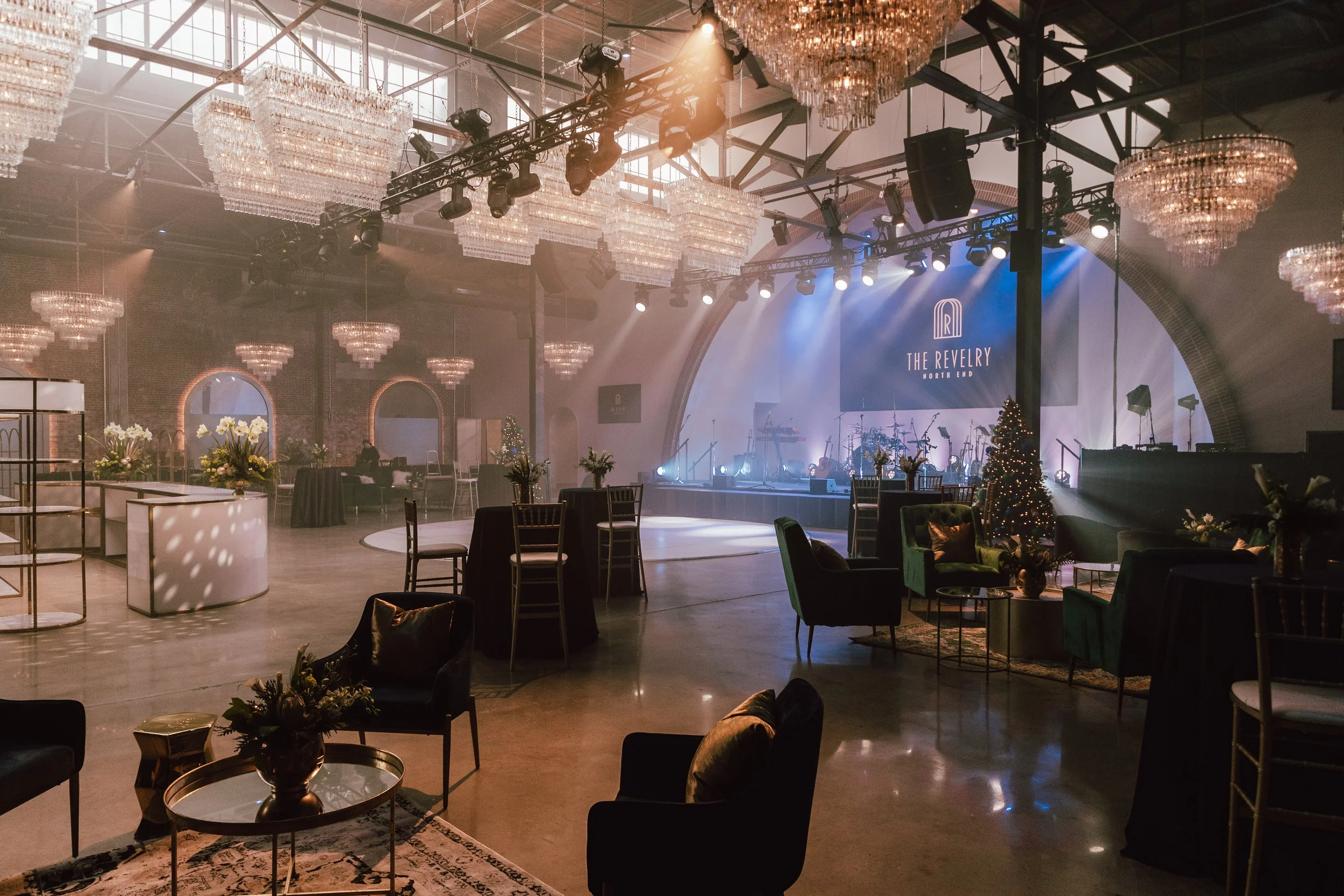 An event space with a stage, musical instruments, and a large screen displaying 'The Revelry North End'. The space has chandeliers, decorated with floral arrangements, a Christmas tree, and elegant furniture, set for a festive gathering.