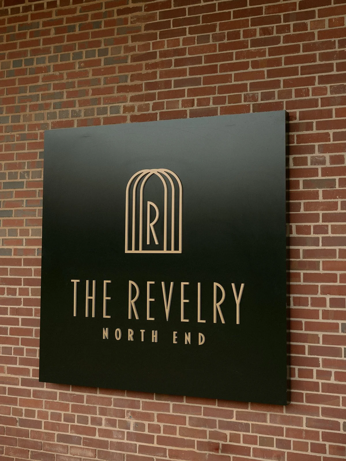 Sign with black background and white lettering that reads 'The Revelry North End' with a stylized letter R logo above the text, mounted on a brick wall.