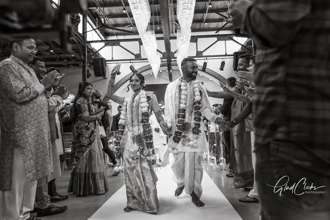 A wedding ceremony with a bride and groom walking down an aisle, holding hands, and surrounded by guests clapping and celebrating indoors.