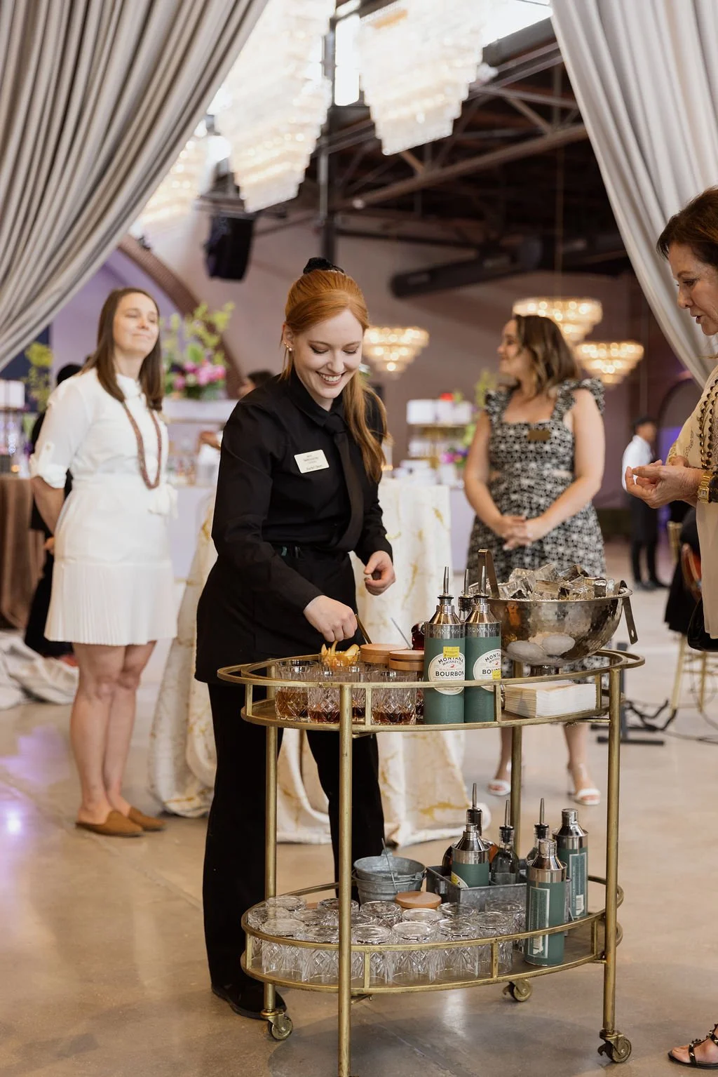 A bartender serving drinks at a social event, with women in the background, a chandelier, and a decorated dance floor.