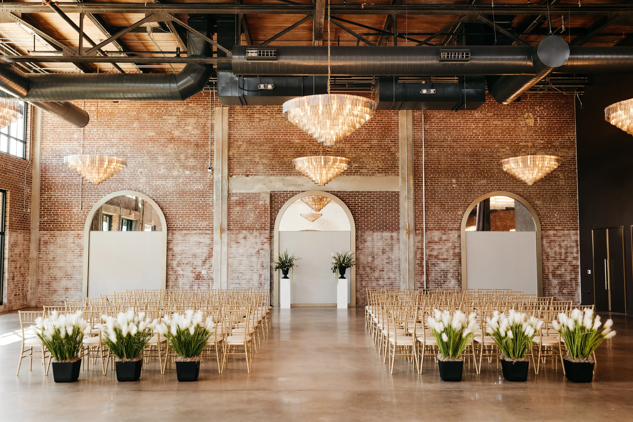 Indoor event space with rows of wooden chairs, potted white flowers, exposed brick walls, large arched mirrors, and elegant chandeliers hanging from a black ceiling with visible ductwork.