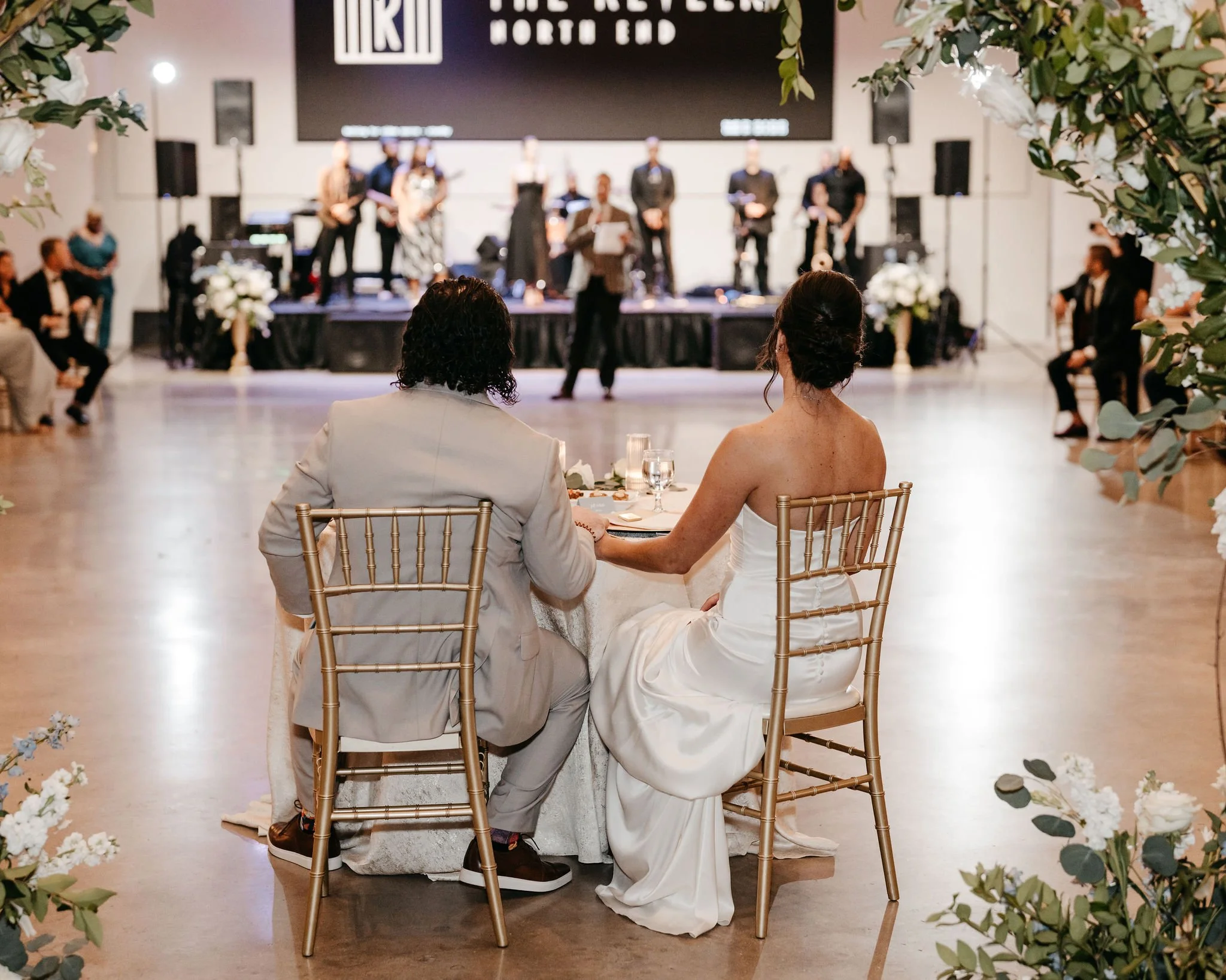 A bride and groom holding hands and sitting at a wedding reception table, facing a live band on stage. The bride is in a white strapless dress, and the groom is in a light-colored suit. The stage backdrop displays a large black and white sign with th