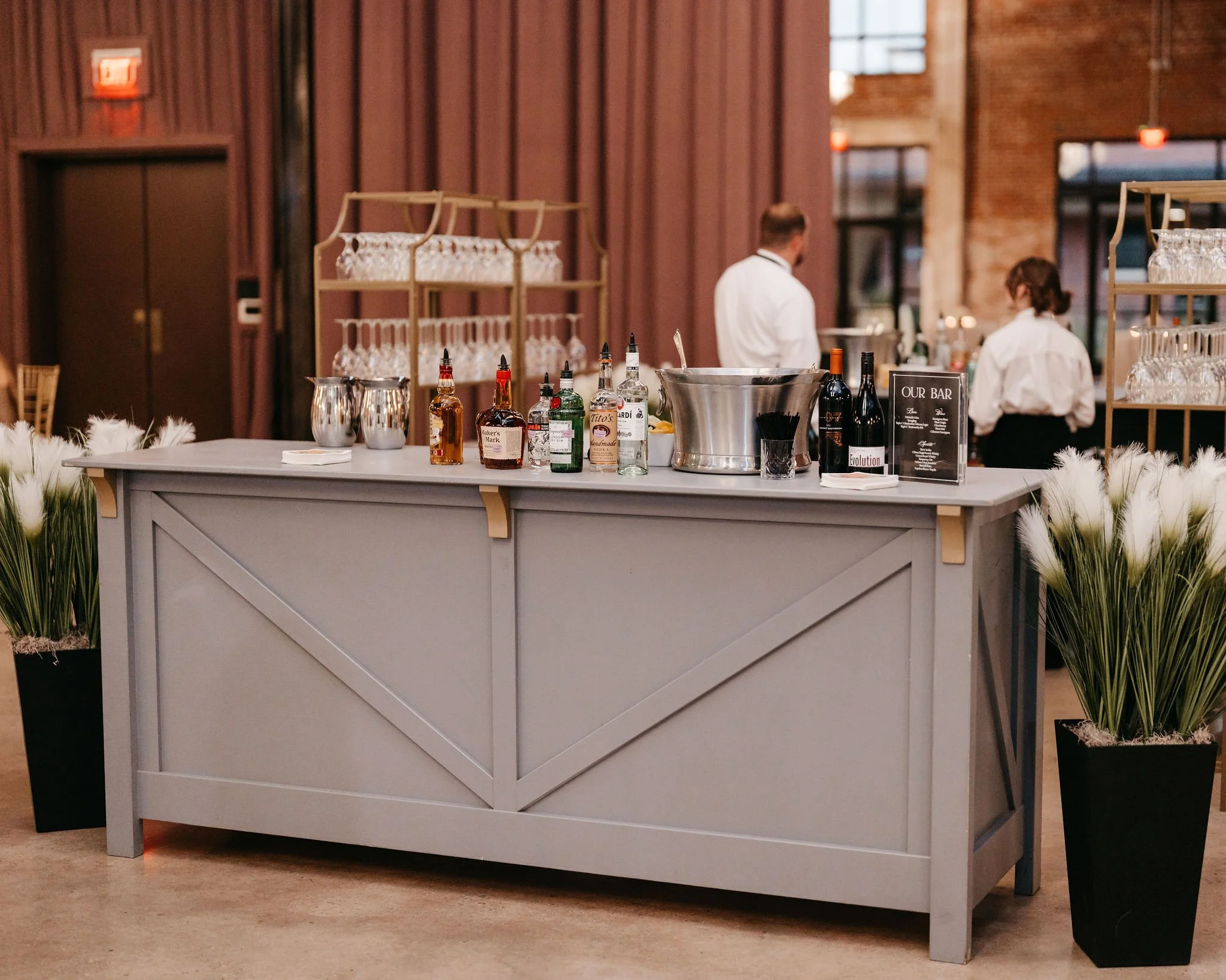 A stylish bar setup with various alcohol bottles, glasses, and silver ice buckets, flanked by two large potted plants with white fluffy flowers in black containers, inside a rustic venue with brick and wood decor.
