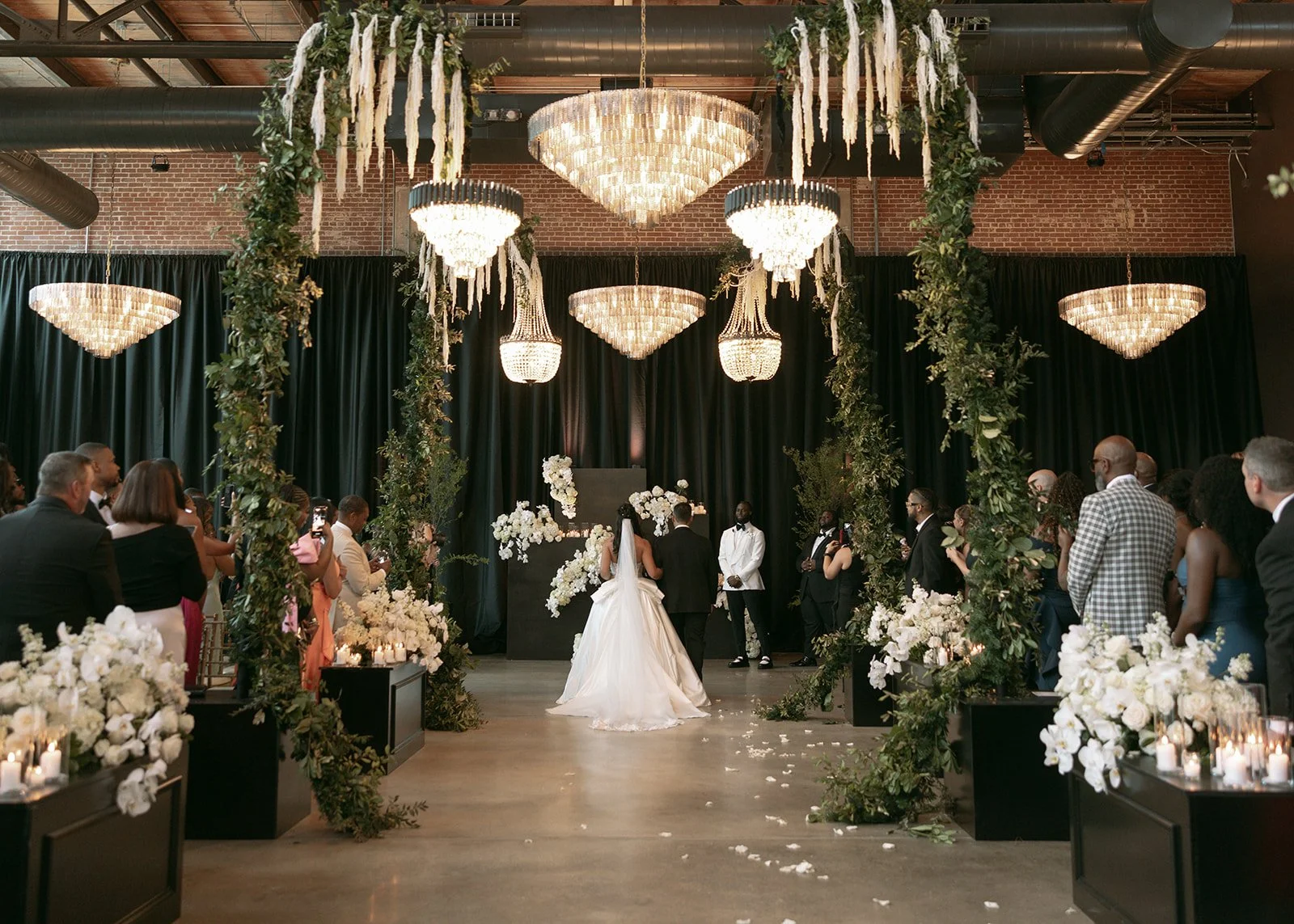 A wedding ceremony with a bride and groom walking down the aisle, surrounded by guests. The venue features elegant chandeliers, black curtains, and floral decorations with white flowers and greenery.