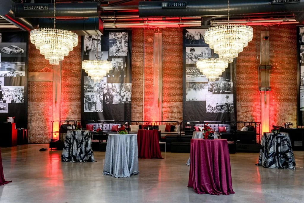 Elegant event space with high ceilings, brick walls, multiple chandeliers, and black-and-white historical photographs on the walls. Several cocktail tables with colorful tablecloths and floral centerpieces are arranged on a polished wooden floor, wit