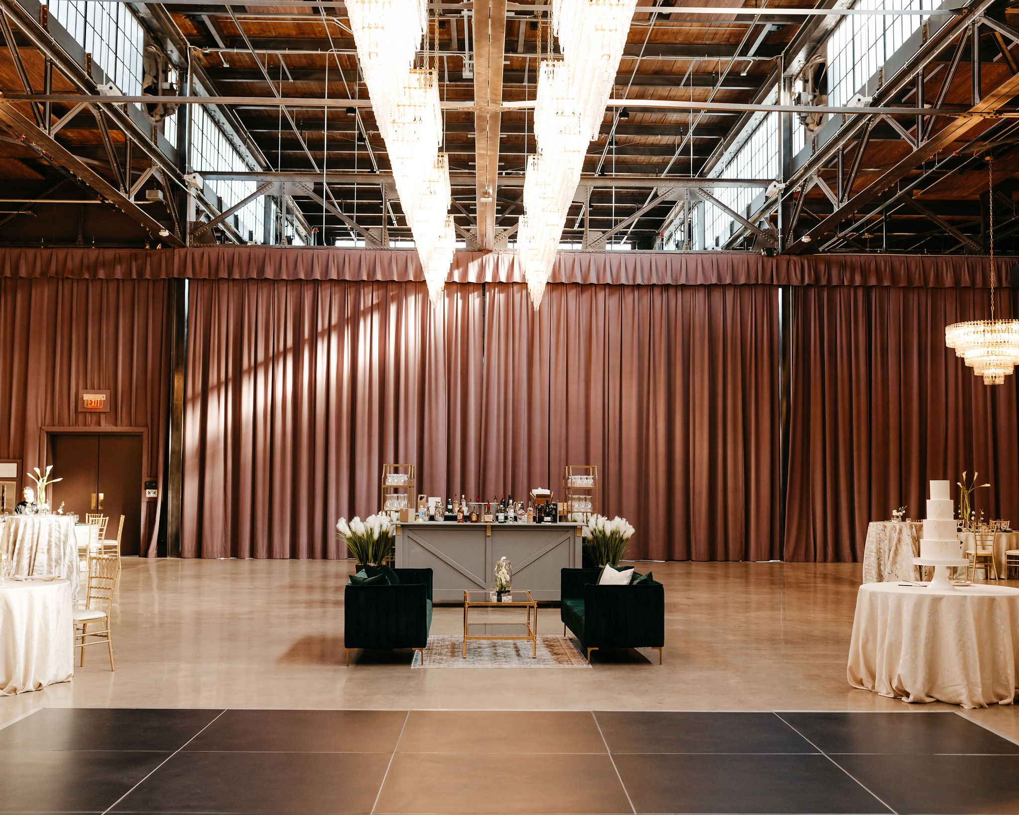 Decorated event space with a large brown curtain, chandeliers, a bar in the center, and tables with floral arrangements and decorations.