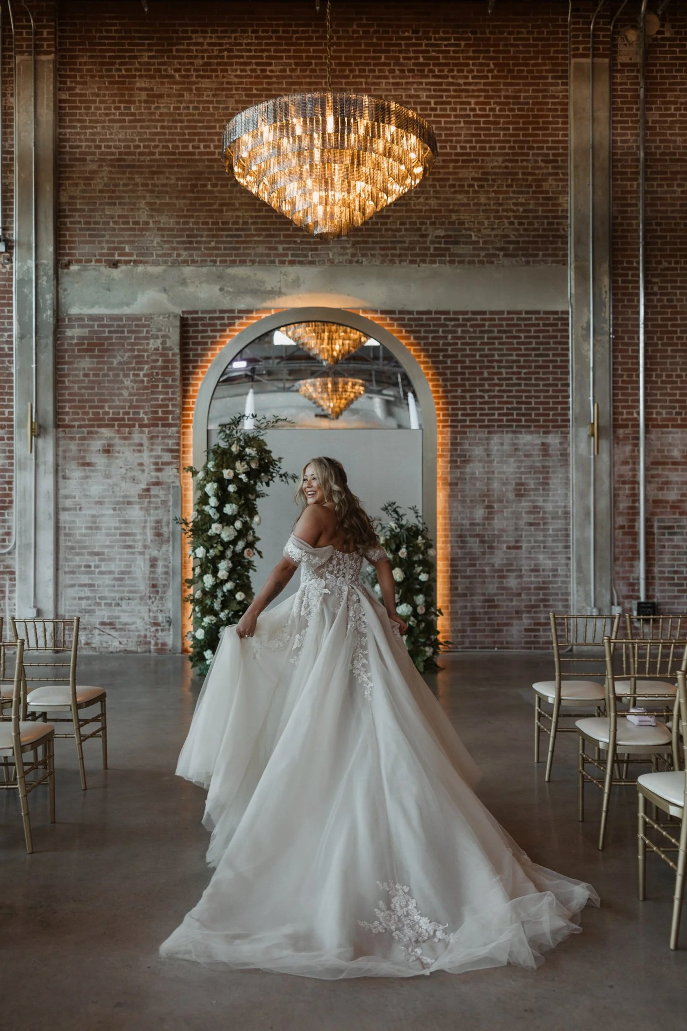 A woman in a wedding dress standing in an industrial-style venue with brick walls, floral arrangements, a large mirror, and elegant gold chairs.