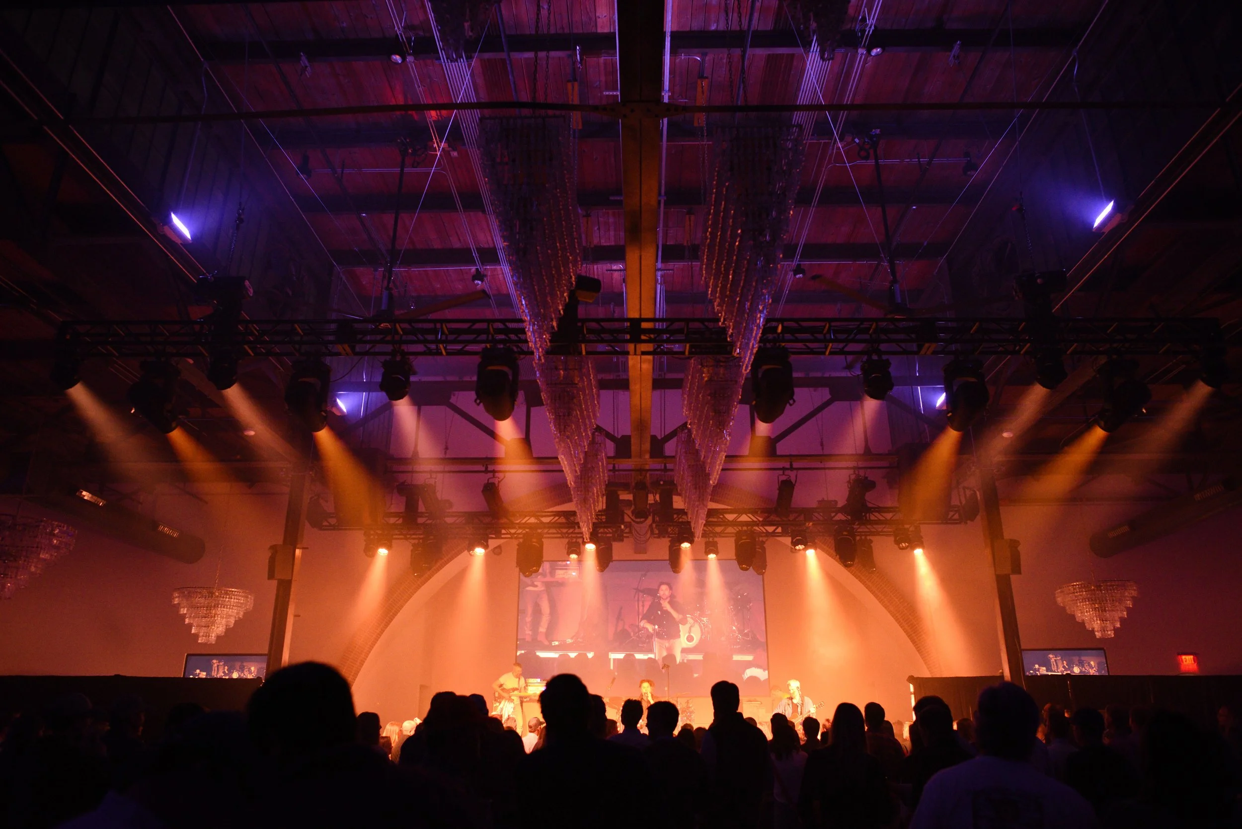 Live music concert with performers on stage and audience watching, illuminated by orange and purple stage lights in a large indoor venue.