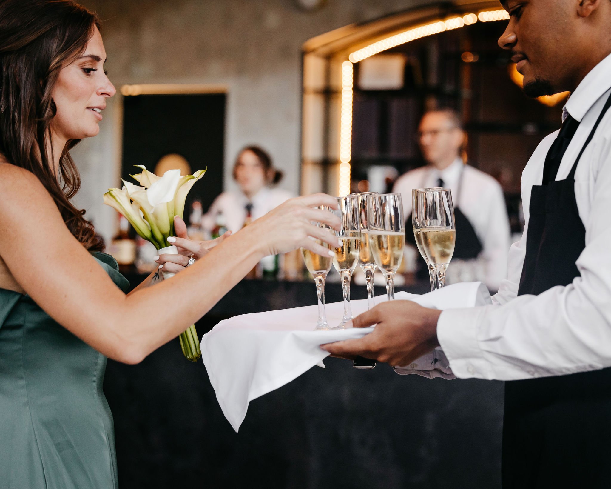 A woman in a green dress receiving a tray of champagne glasses from a server at a celebration or event.