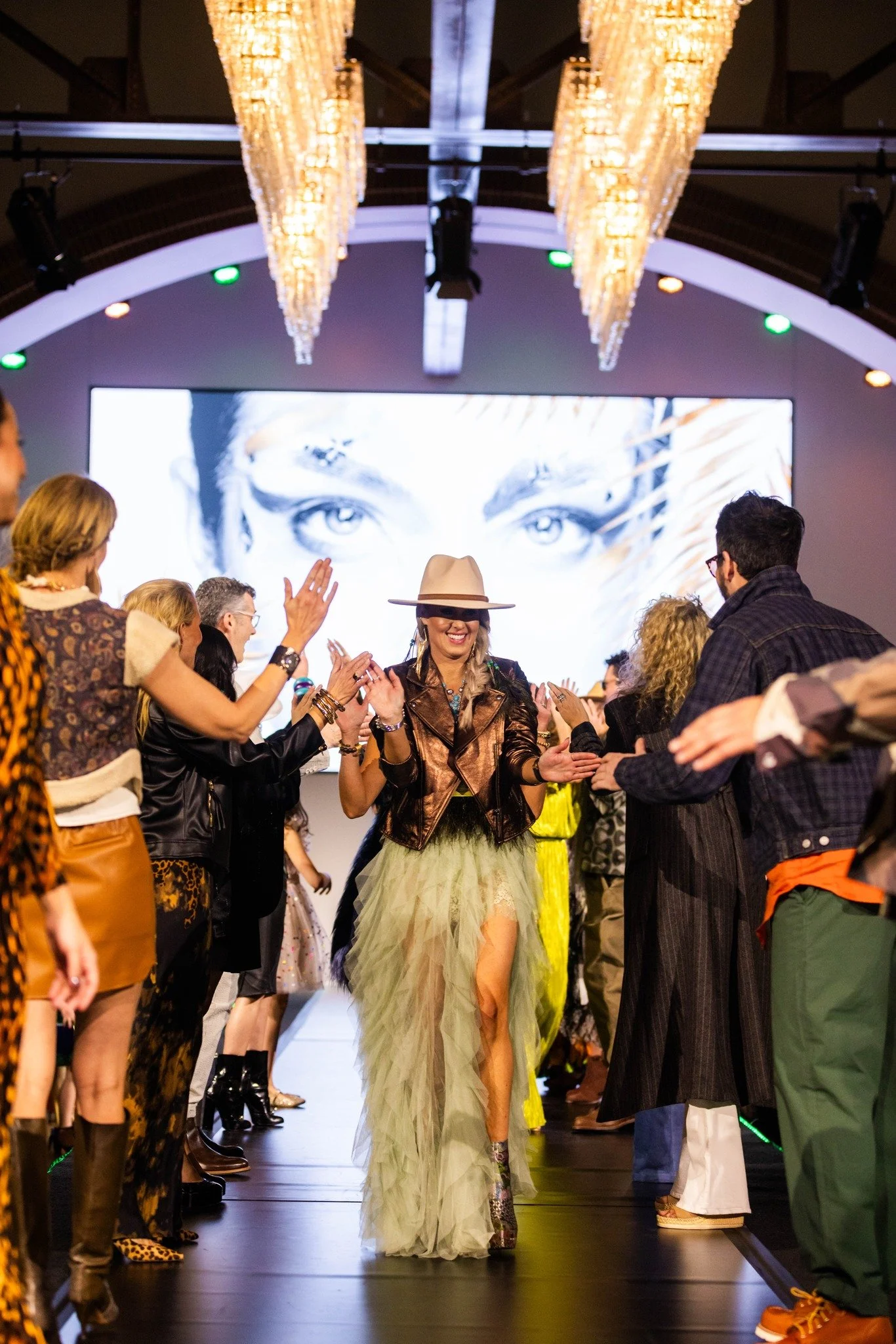 A woman wearing a tan hat, a metallic bronze jacket, and a flowing yellow-green gown with a high slit, walking on a runway at a fashion show surrounded by clapping spectators.