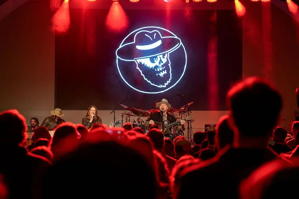 A live concert featuring a band on stage with a large neon skull with a beard and hat in the background, and an audience watching.