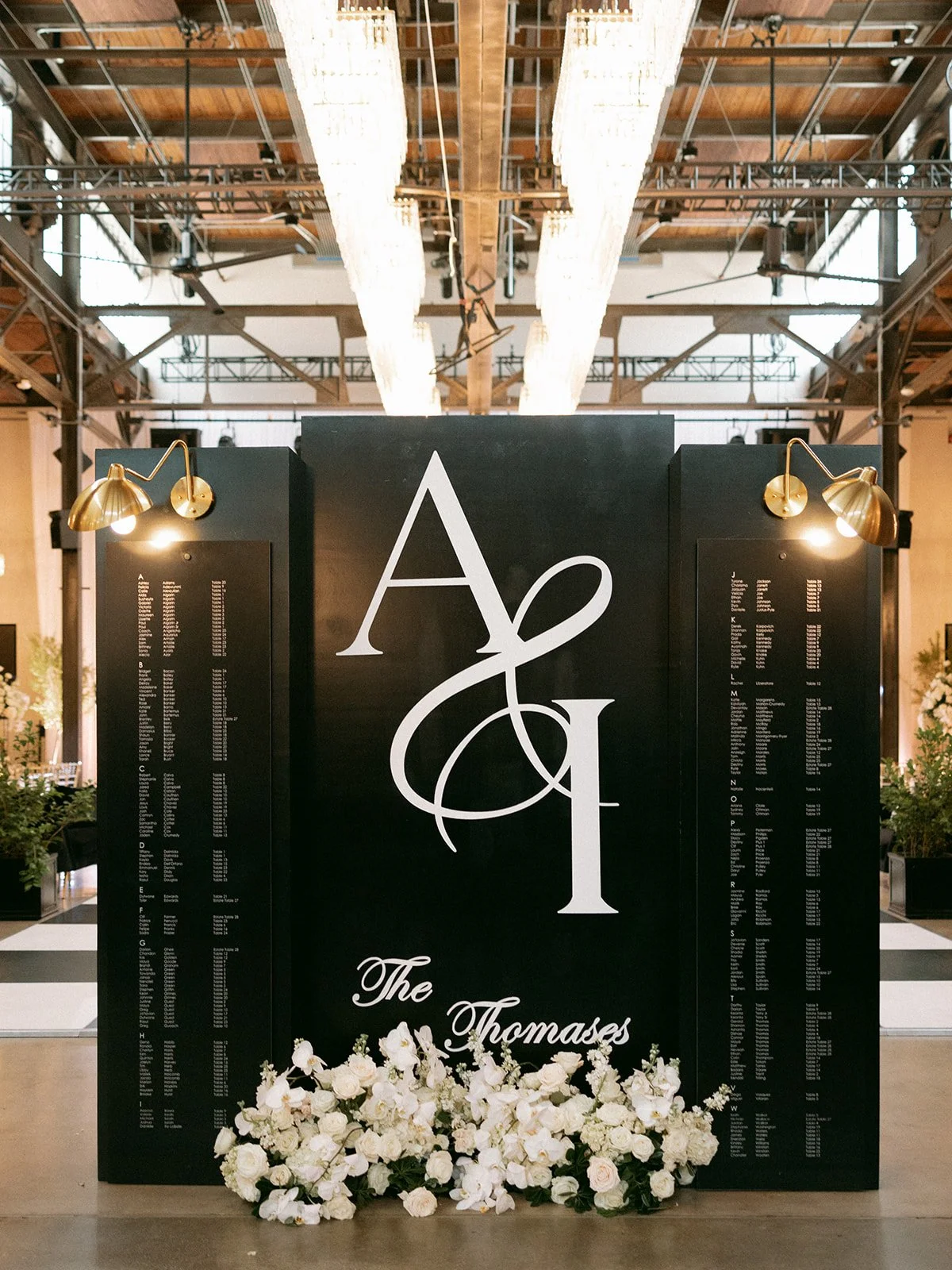 A black wedding seating chart display with large white initials 'A & J' and the words 'The Thomases' written below, decorated with white flowers at the base, flanked by illuminated brass wall sconces, in an elegant hall with high ceilings, chandelier