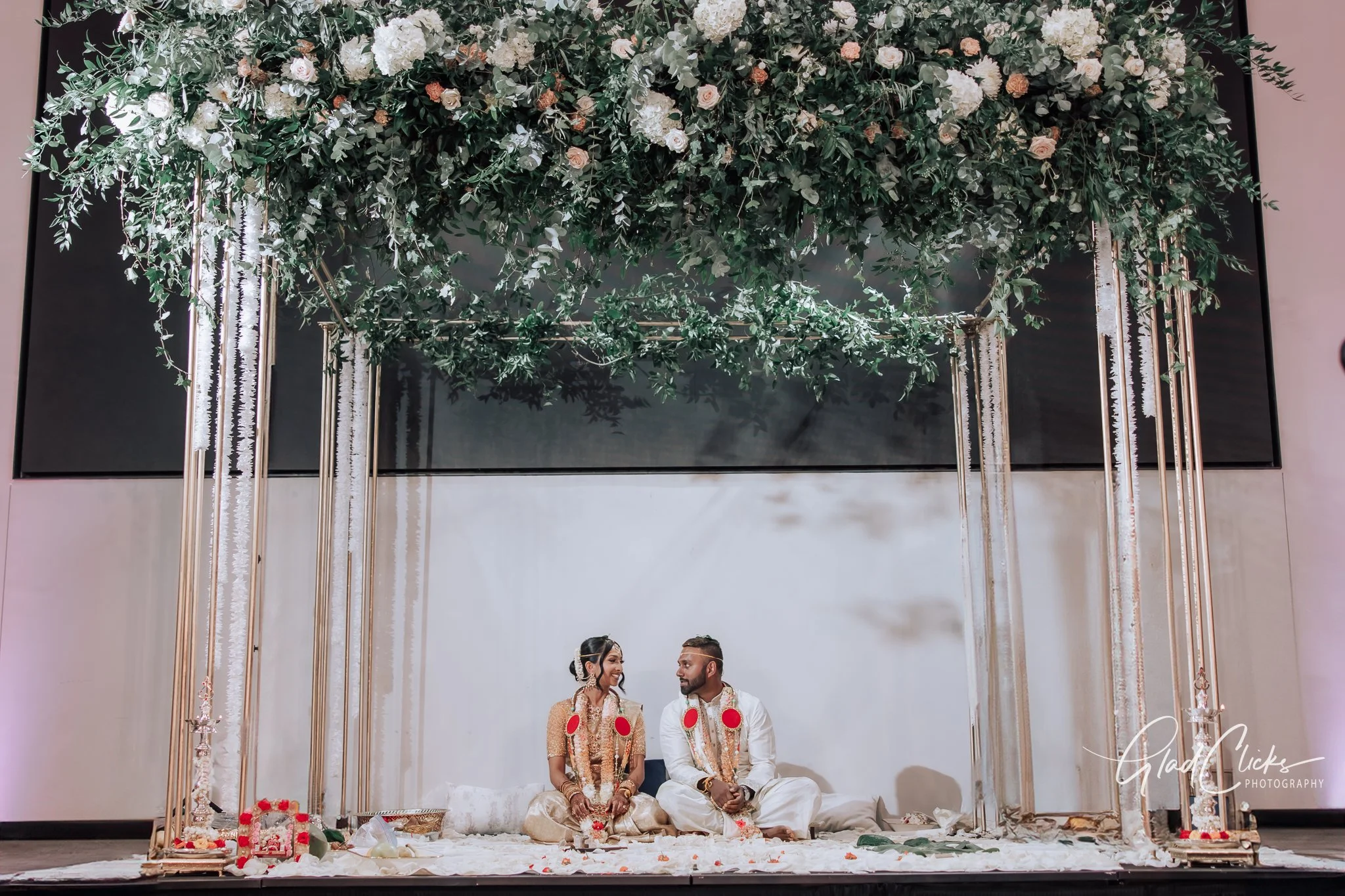 A traditional Indian wedding ceremony with a bride and groom sitting on the floor under a decorated floral canopy. The canopy is adorned with greenery and white and peach flowers. The couple is dressed in traditional Indian attire, with the bride in 
