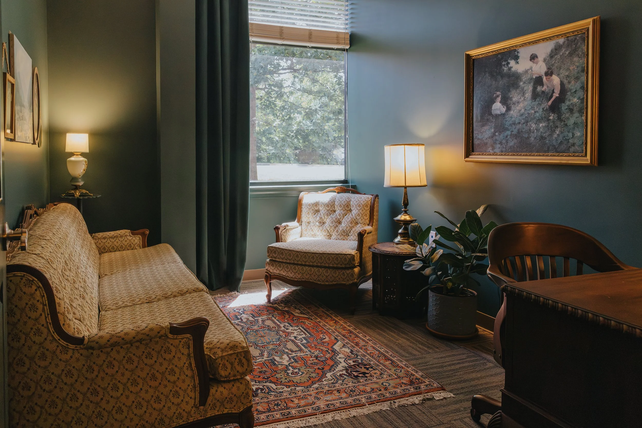 A couch, desk, and chair in a moody, Victorian apothecary-style forest green room with a window.