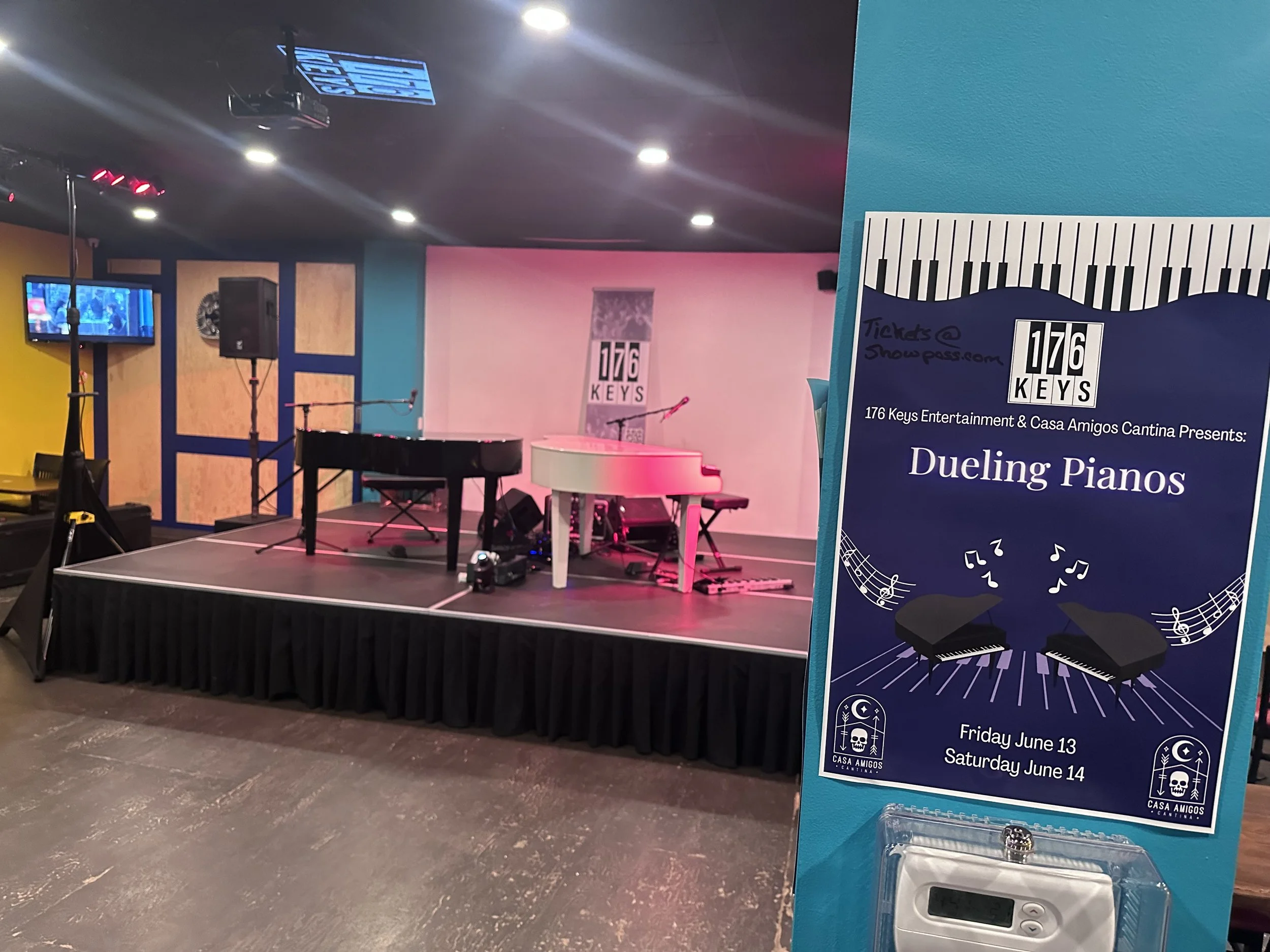 Empty stage with two pianos, one black and one white, set for a musical performance at Casa Amigos Cantina with colorful wall lighting. A promotional flyer for the event 'Dueling Pianos' is displayed in the foreground.