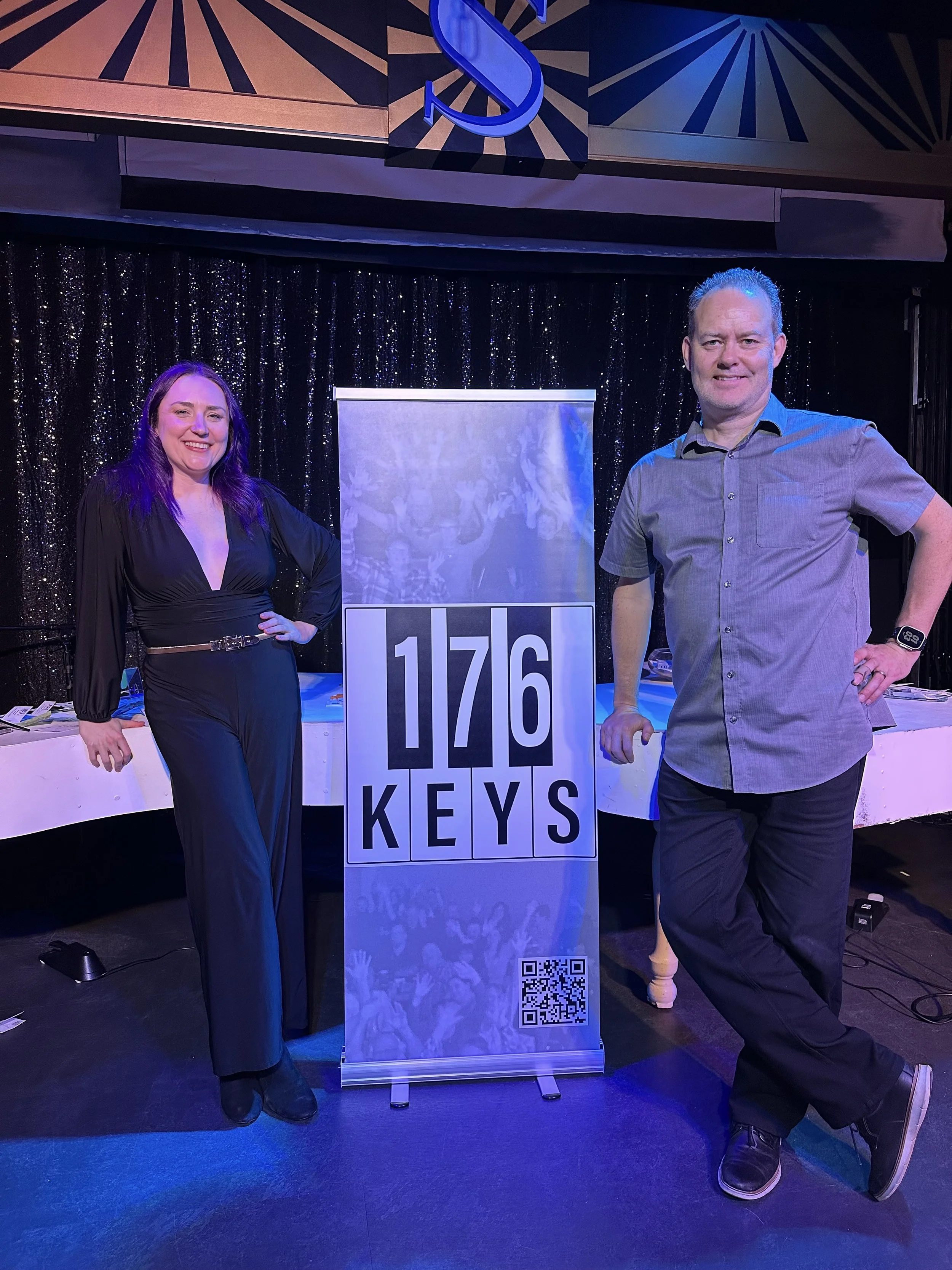 Two people standing next to a sign that reads '1776 KEYS' with a QR code at the bottom, in front of a black sequin backdrop and a decorated stage.