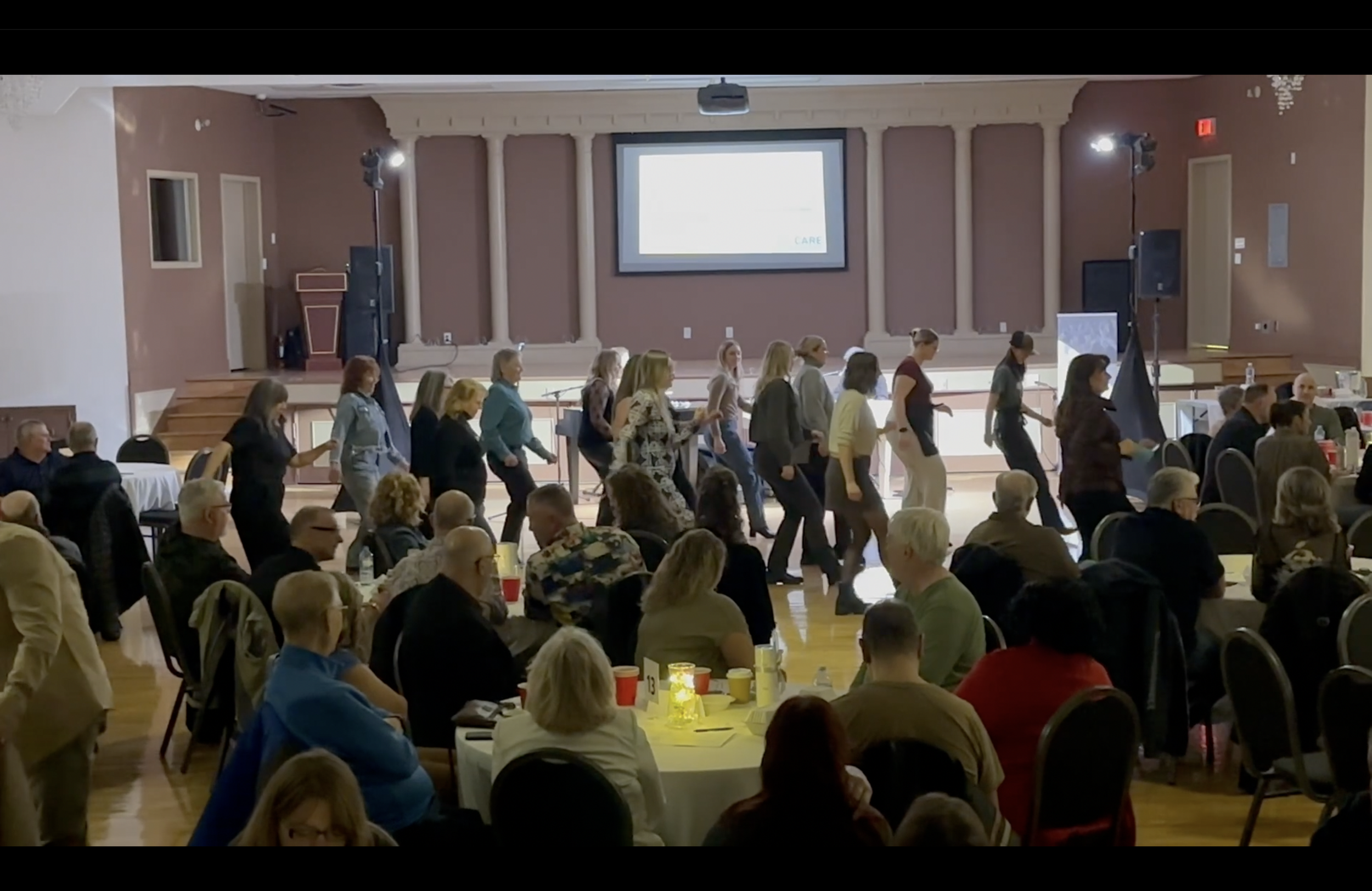 People line dancing at an event in a large auditorium with round tables, audience seated, a stage with a large screen in the background.