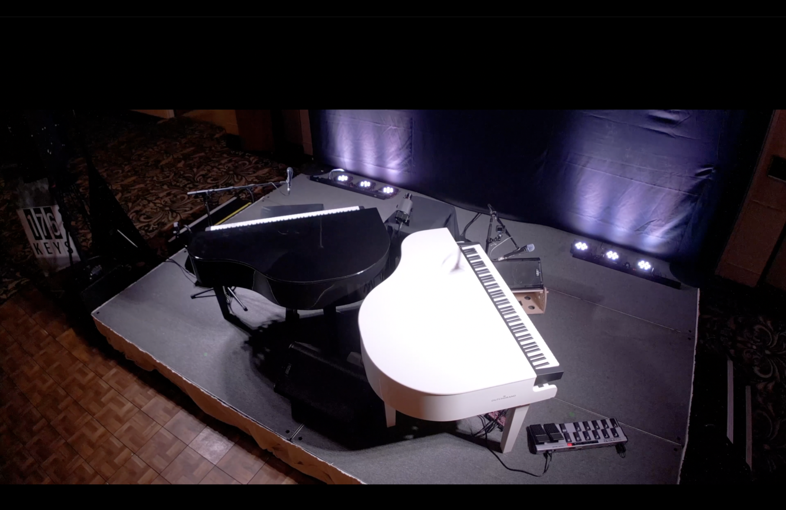 A stage setup with a black grand piano, a white digital piano, microphones, and stage lights in a dimly lit room.