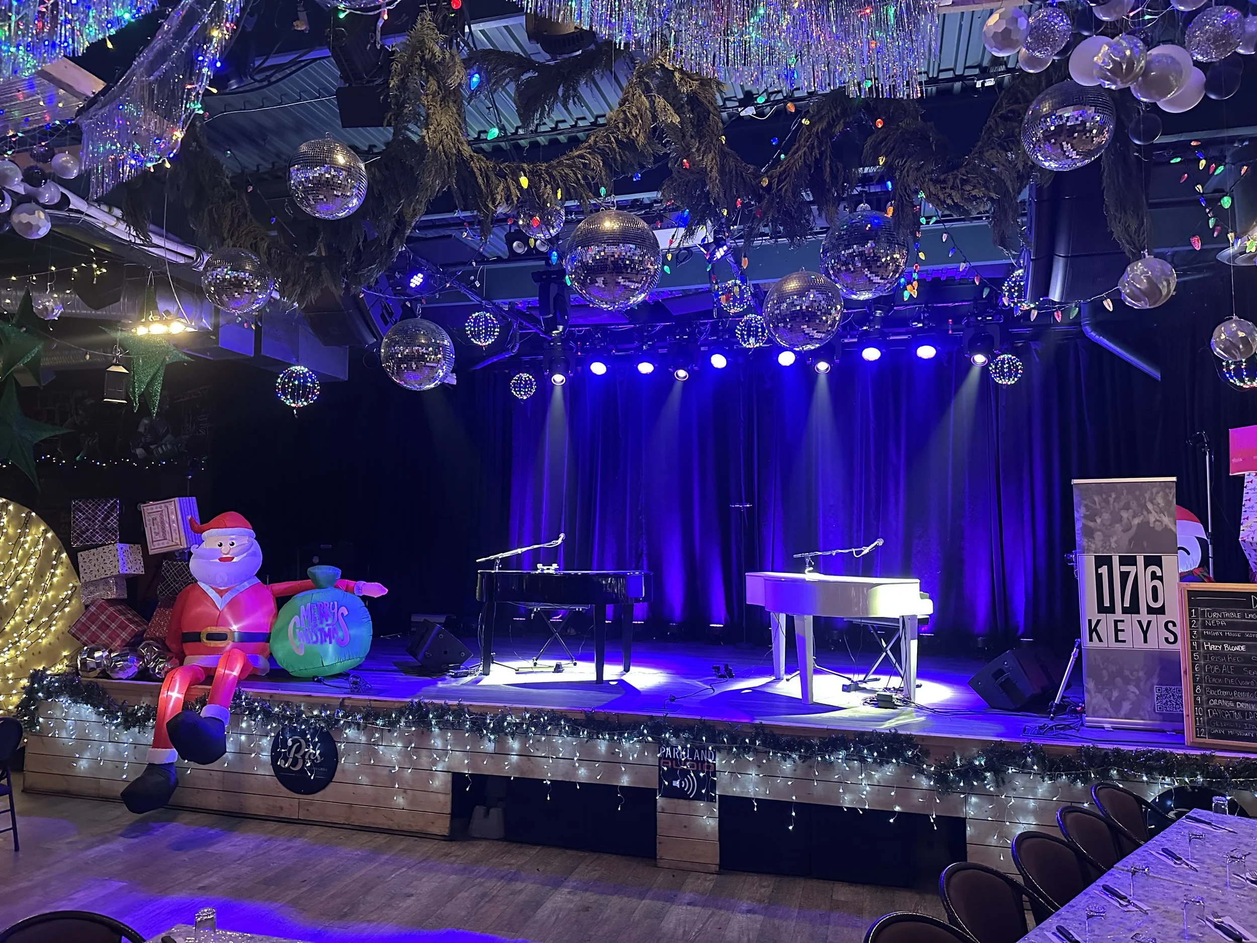 Stage decorated with Christmas ornaments, including hanging disco balls and tinsel, with a large inflatable Santa figure and colorful decorations. The stage has two pianos and a black curtain backdrop, with seating and tables in front.