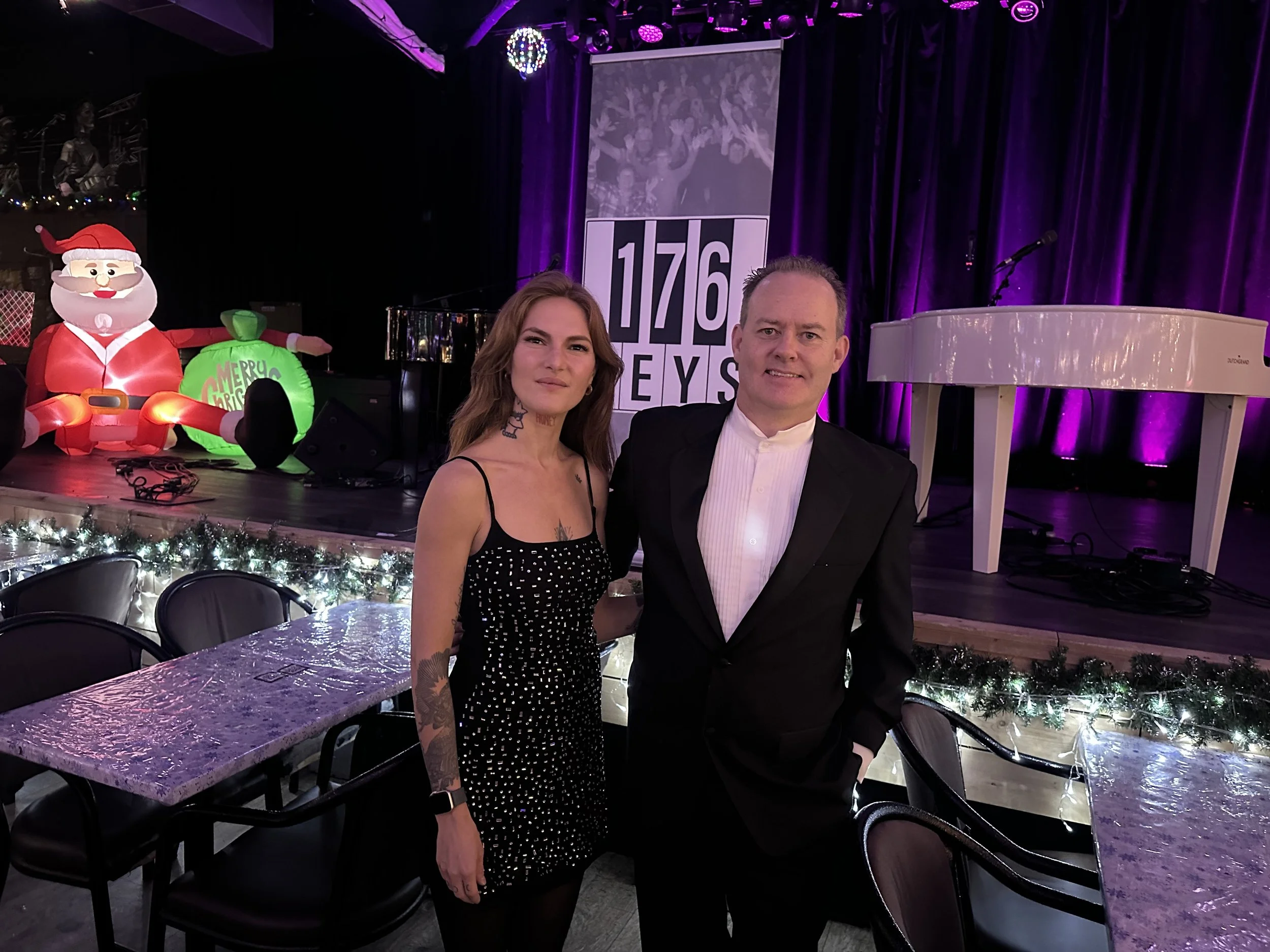 A woman in a black dress with white polka dots and tattoos on her arm stands next to a man in a tuxedo, both smiling. They are in a decorated event space with a stage in the background featuring inflatable Santa and Christmas decorations, purple ligh