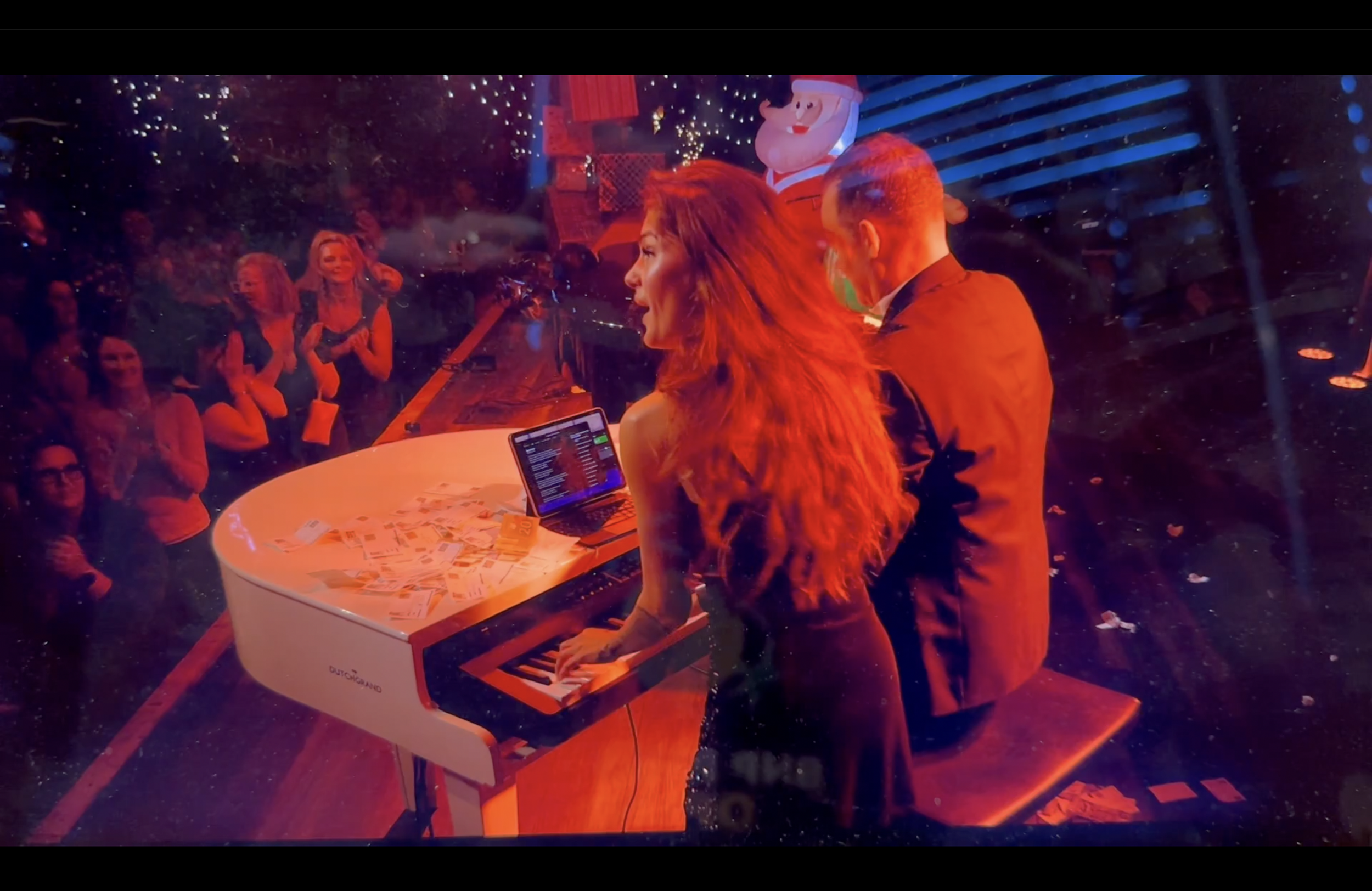 A woman and a man perform on a white grand piano during a festive holiday event, with a crowd of spectators watching. There are Christmas decorations, including a Santa Claus figure and an inflatable Christmas elf, in the background.