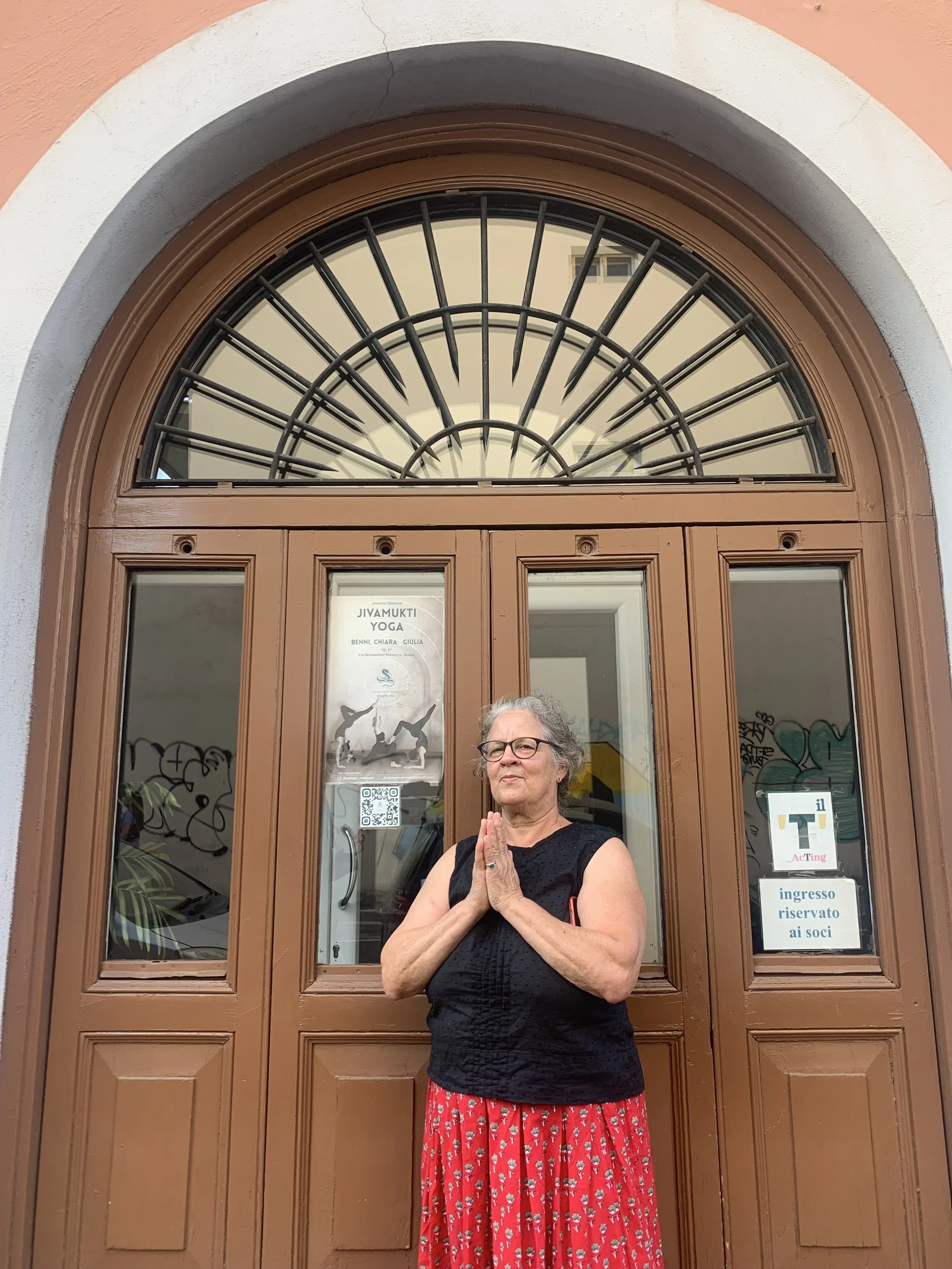 An elderly woman with glasses and gray hair, wearing a black sleeveless top and a red skirt with floral pattern, stands with her hands pressed together in a prayer-like gesture in front of a brown wooden door with an ornate arched window above it. The door has signs and posters, including one for yoga classes and another indicating the entrance is reserved for members.