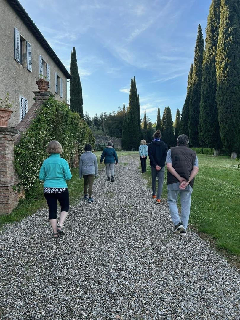A group of people walking along a gravel pathway in a scenic outdoor setting with tall cypress trees and a stone building with shuttered windows on the left.